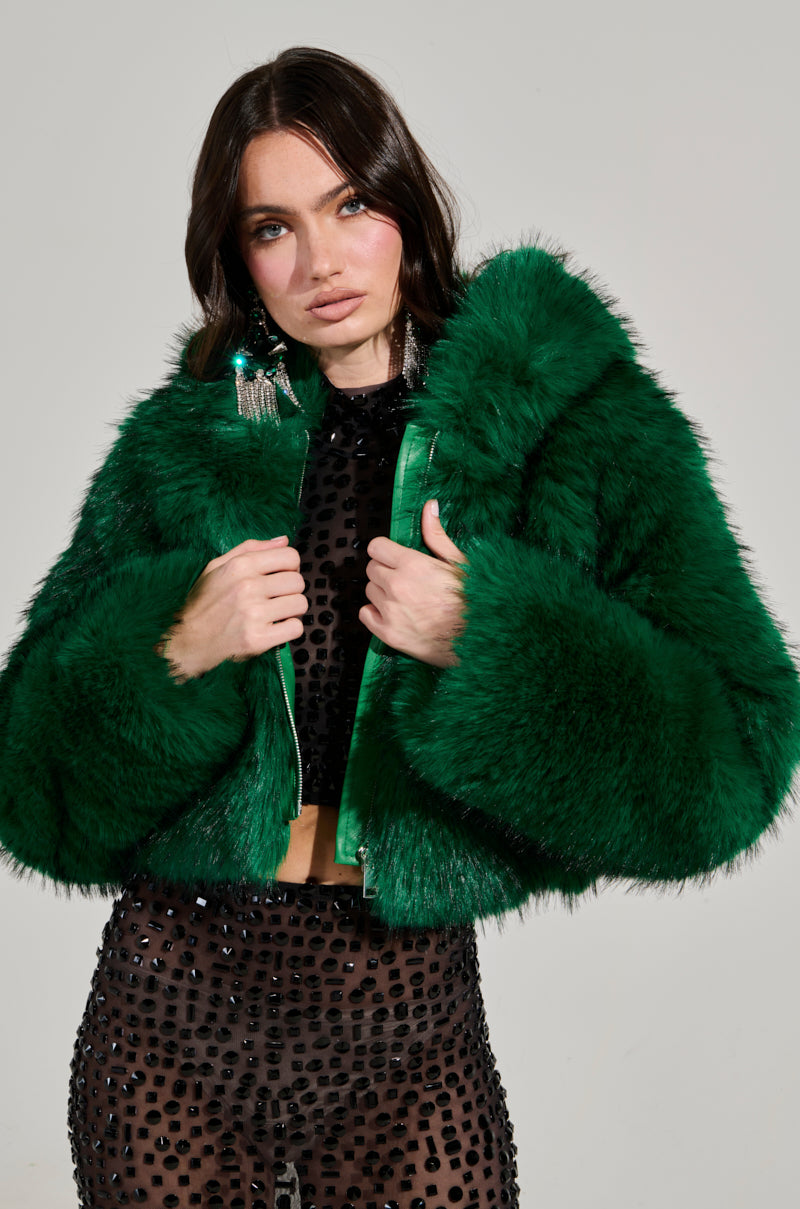 A person wearing the LUCKY HOODED FUR BOMBER in a vibrant green hue over a sheer black top with textured patterns stands against a plain background. They have long dark hair and wear large, dangling earrings.
