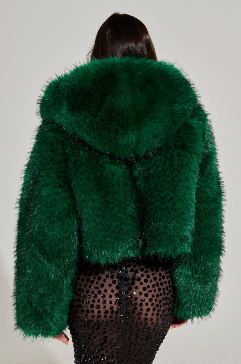 Wearing the LUCKY HOODED FUR BOMBER, she showcases her back in a green faux fur jacket, perfectly paired with a sheer black skirt embellished with circular designs, all elegantly framed by a neutral backdrop that enhances her look.