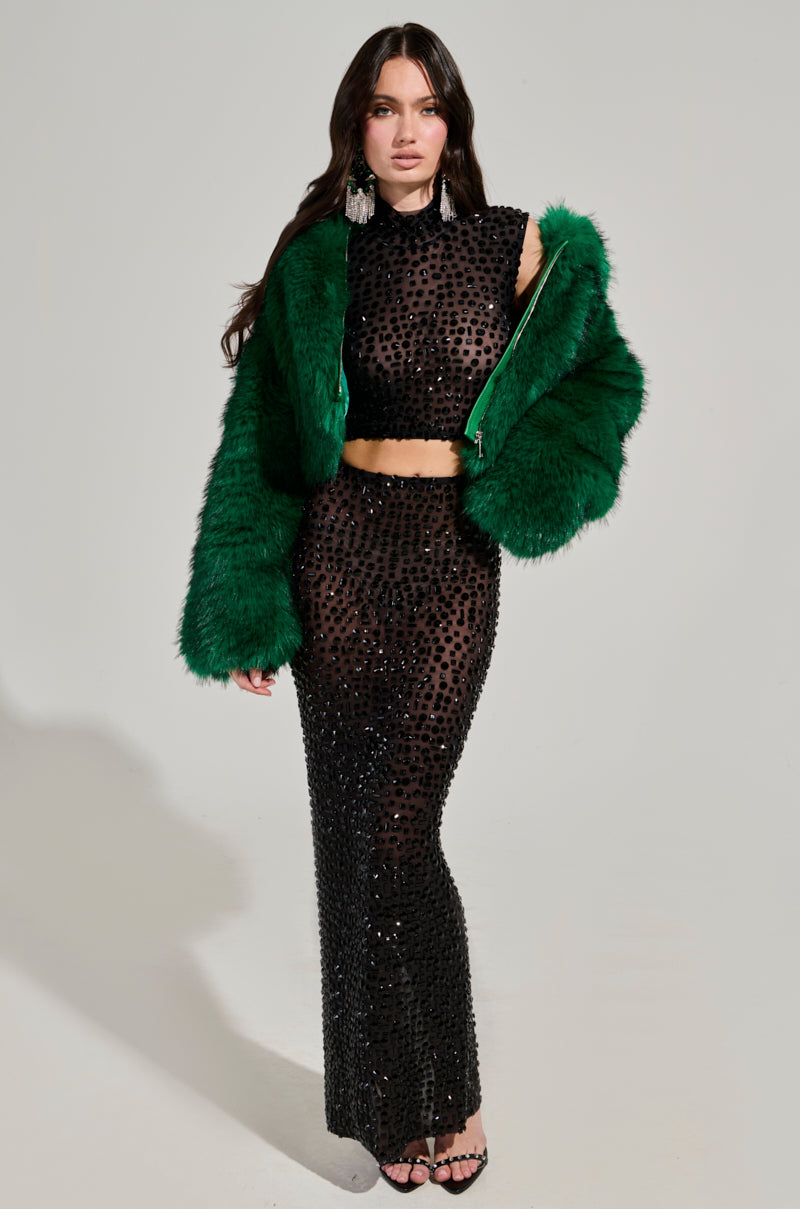 Dressed to impress, a woman exudes confidence in a black, sheer, beaded dress paired with the striking LUCKY HOODED FUR BOMBER. Her long hair cascades elegantly as she stands tall in black high-heeled sandals against a plain white background.