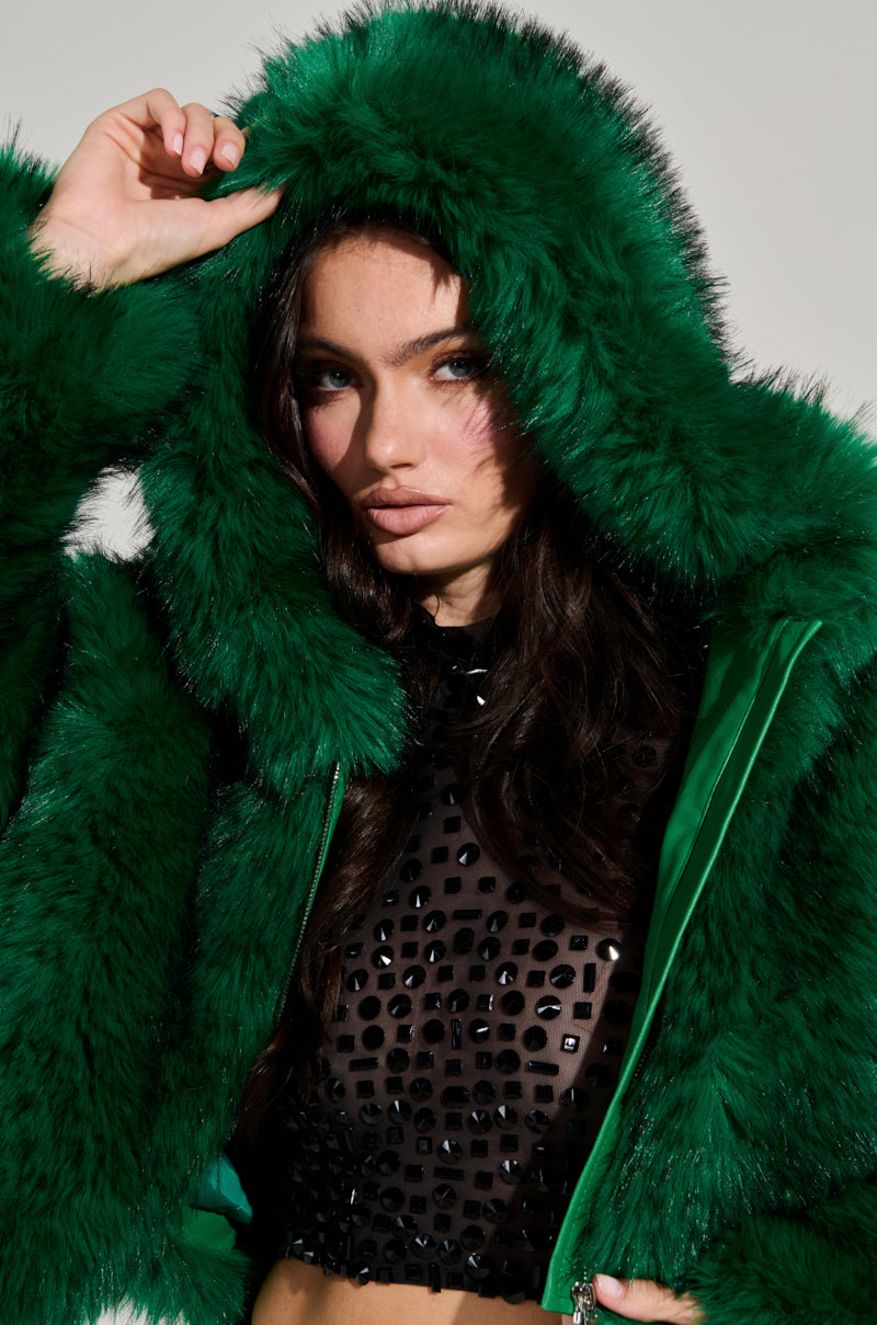 A person wearing the LUCKY HOODED FUR BOMBER in vibrant green with a black sequined top underneath gazes directly at the camera, holding the hood with one hand. The neutral background accentuates the stunning outfit.