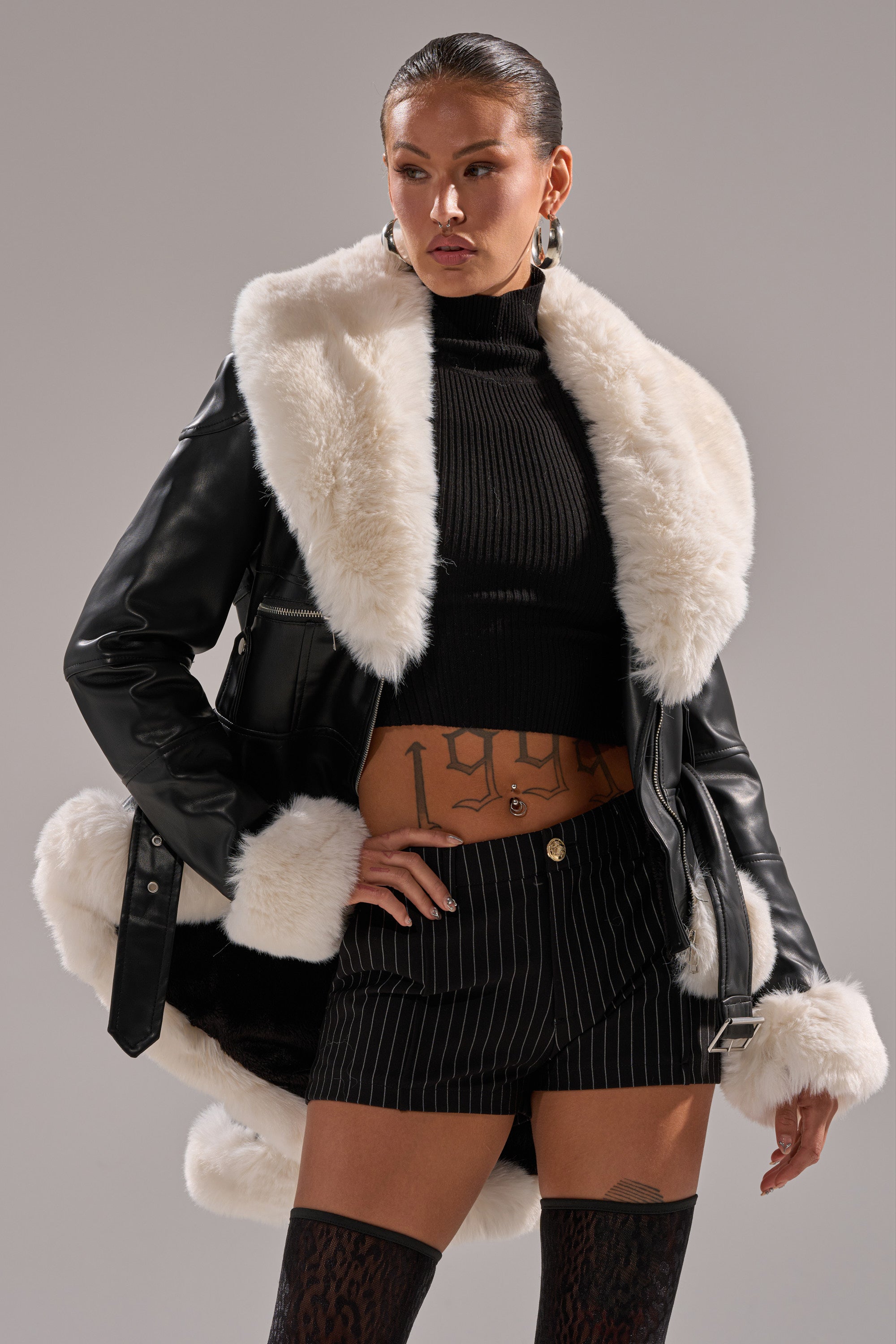 A woman stands with one hand on her hip, looking to the side, wearing the BABY IT'S COLD OUTSIDE FUR TRIM MOTO jacket with a black turtleneck crop top, pinstripe shorts, thigh-high stockings, and hoop earrings.