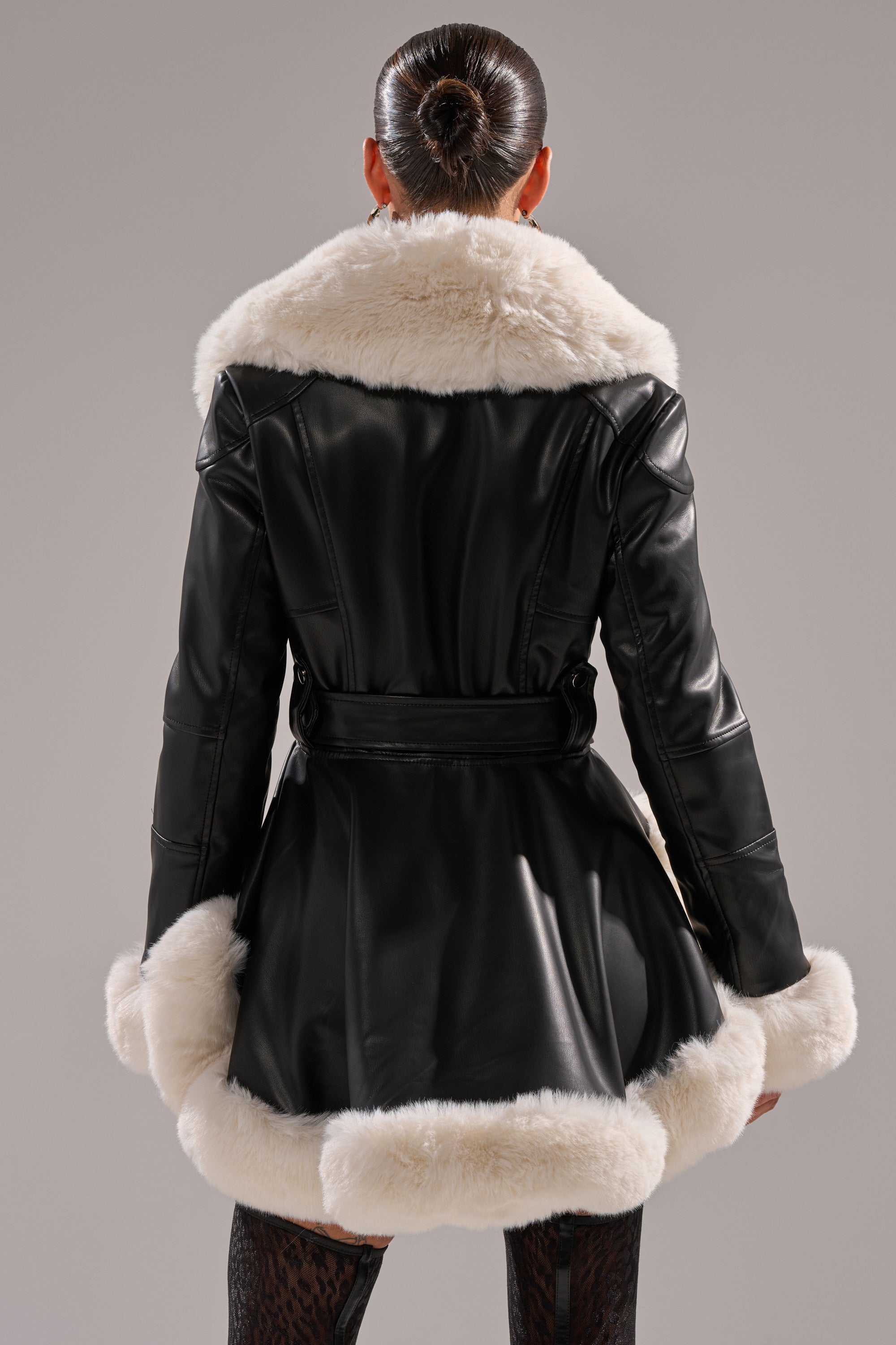 A person with a sleek bun is seen from the back wearing the BABY IT'S COLD OUTSIDE FUR TRIM MOTO, a black faux leather jacket with white fur trim on the collar, cuffs, and hem, set against a plain background.