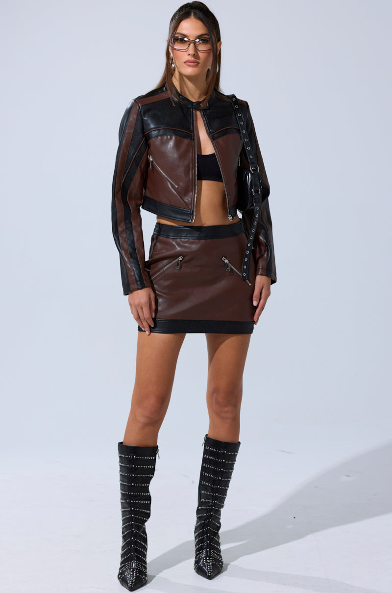 A woman stands against a plain light background wearing the NO PICTURES PLEASE CROPPED MOTO jacket, matching mini skirt with zip details, black crop top, studded knee-high boots, and sunglasses.