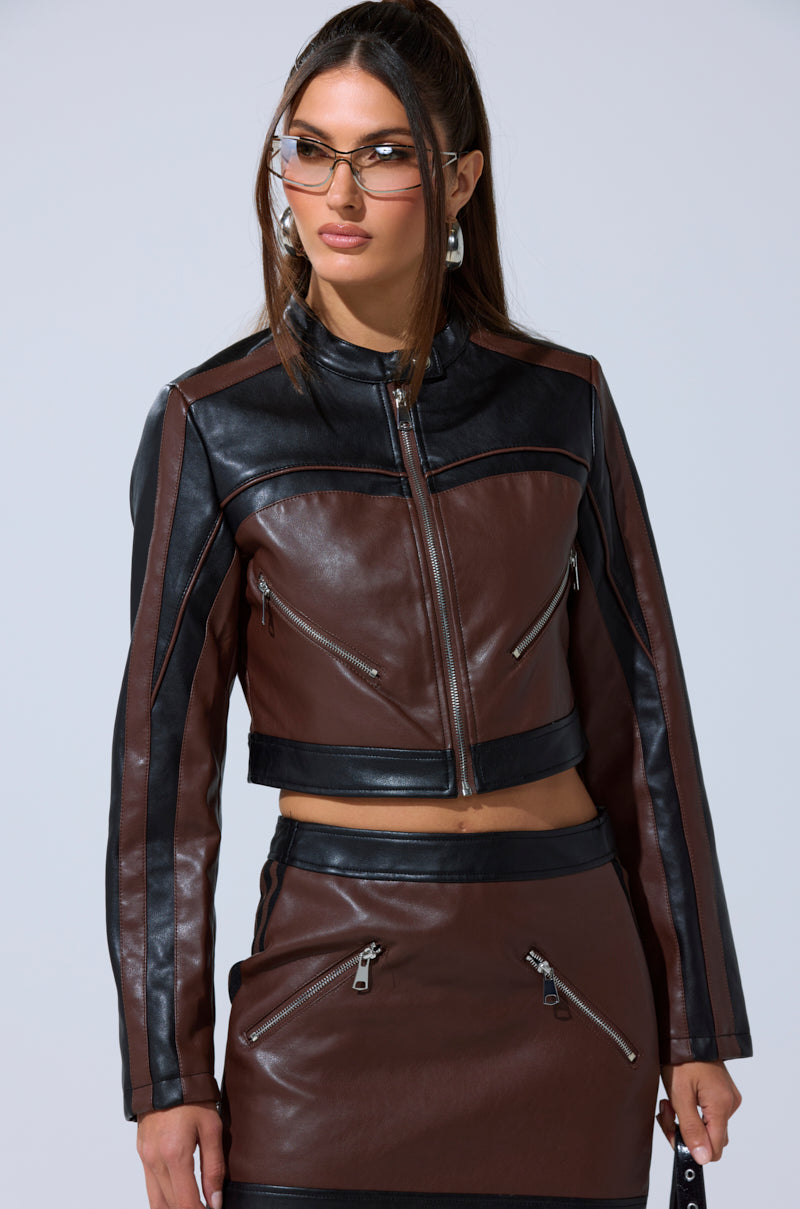 A woman poses confidently in the NO PICTURES PLEASE CROPPED MOTO jacket, paired with a matching skirt, silver hoop earrings, and chic glasses against a plain background.