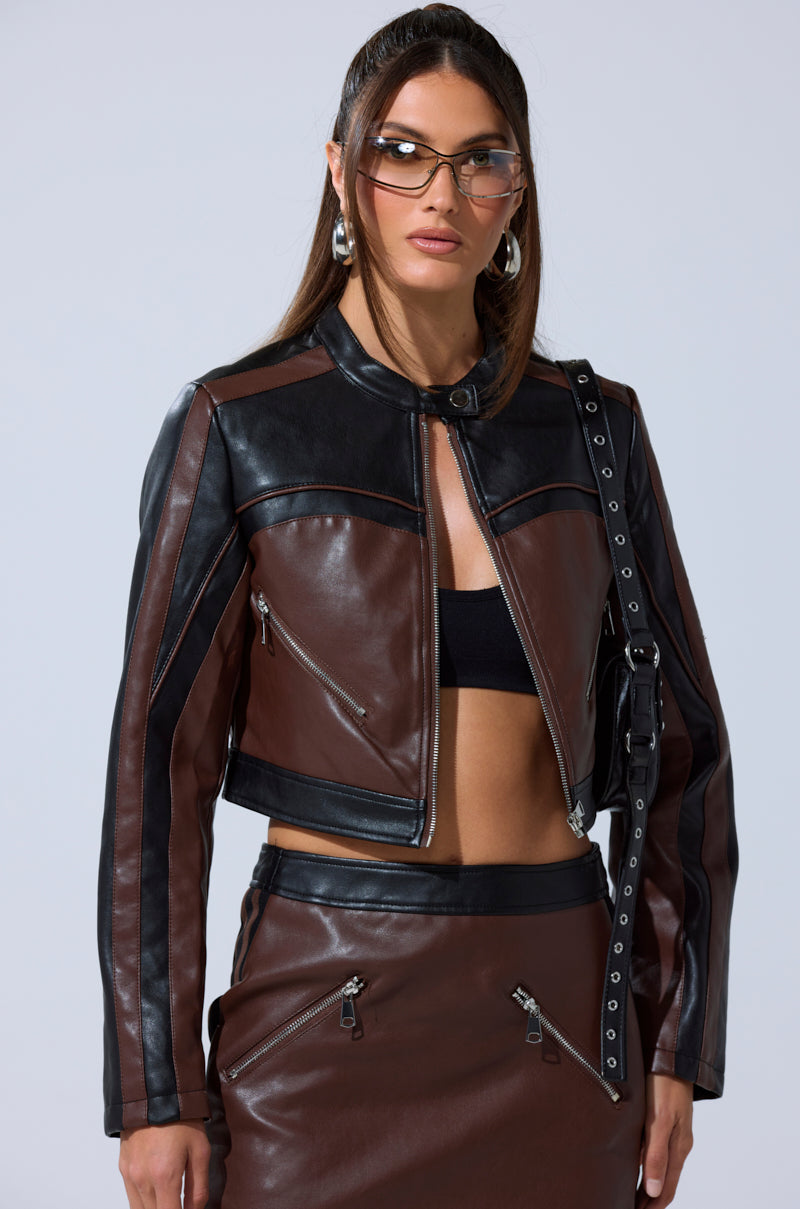 A woman poses against a plain light background wearing the NO PICTURES PLEASE CROPPED MOTO in brown and black with a matching skirt, silver zippers, a black crop top, large hoop earrings, and tinted sunglasses.