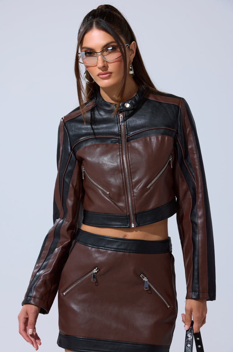 A woman stands against a light background wearing the NO PICTURES PLEASE CROPPED MOTO jacket with zipper details, matching mini skirt, angular glasses, and holding a small bag.