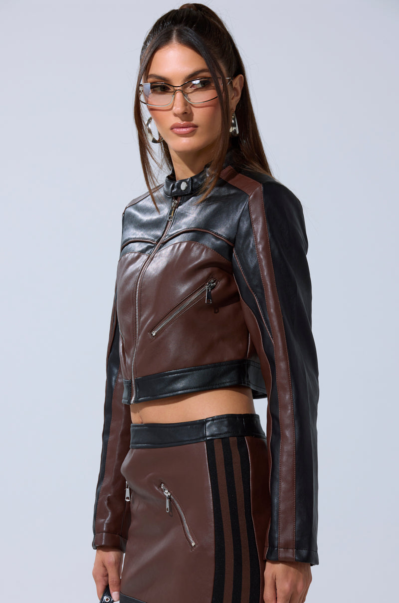 A woman models the NO PICTURES PLEASE CROPPED MOTO jacket and matching skirt with zippered pockets, styled with clear glasses and hoop earrings, standing against a plain light gray background.