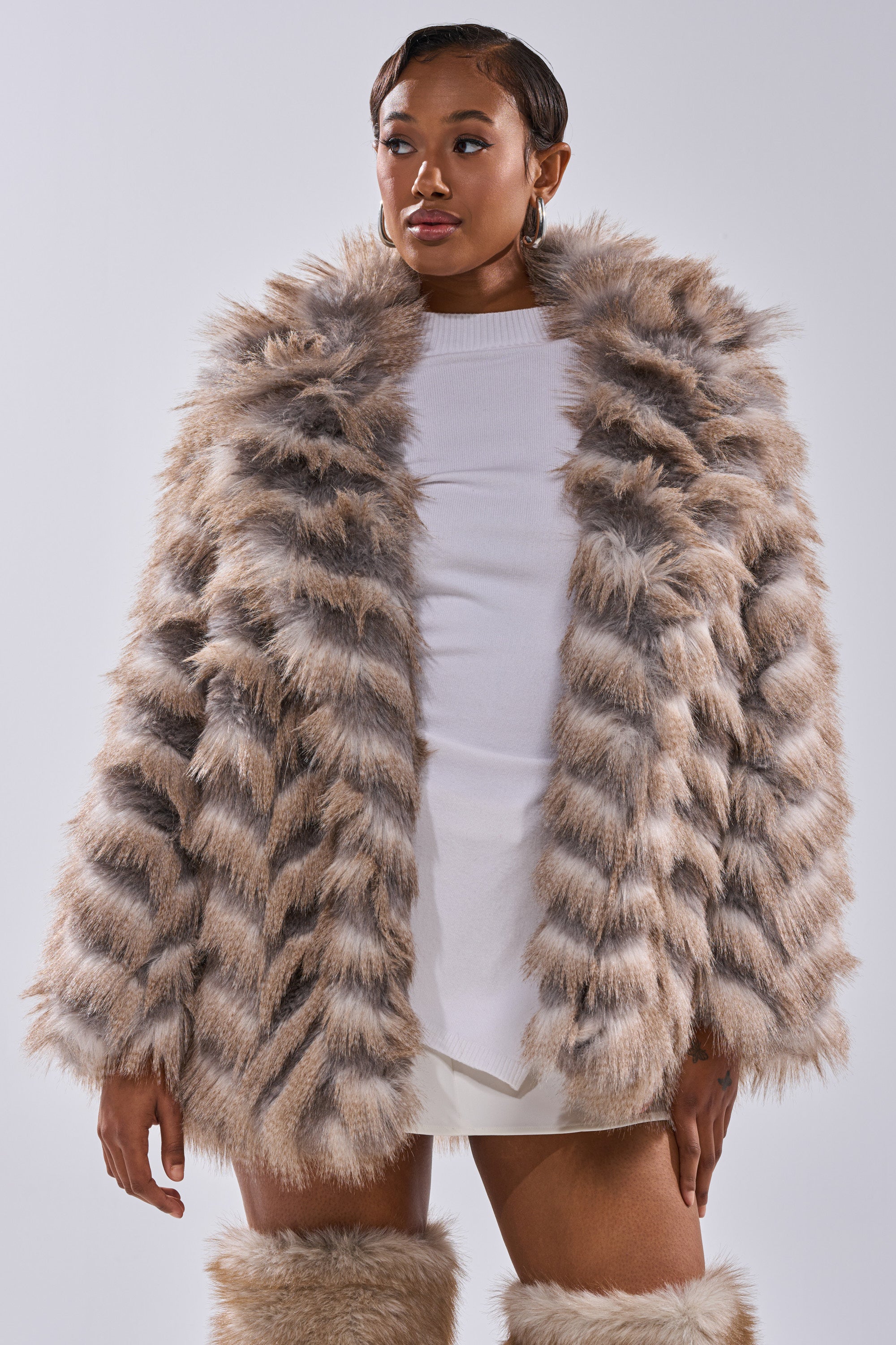 A woman models the SHOWBIZ BABY FAUX FUR coat with a beige and gray chevron print over a white dress and matching furry boots, standing against a plain light background.