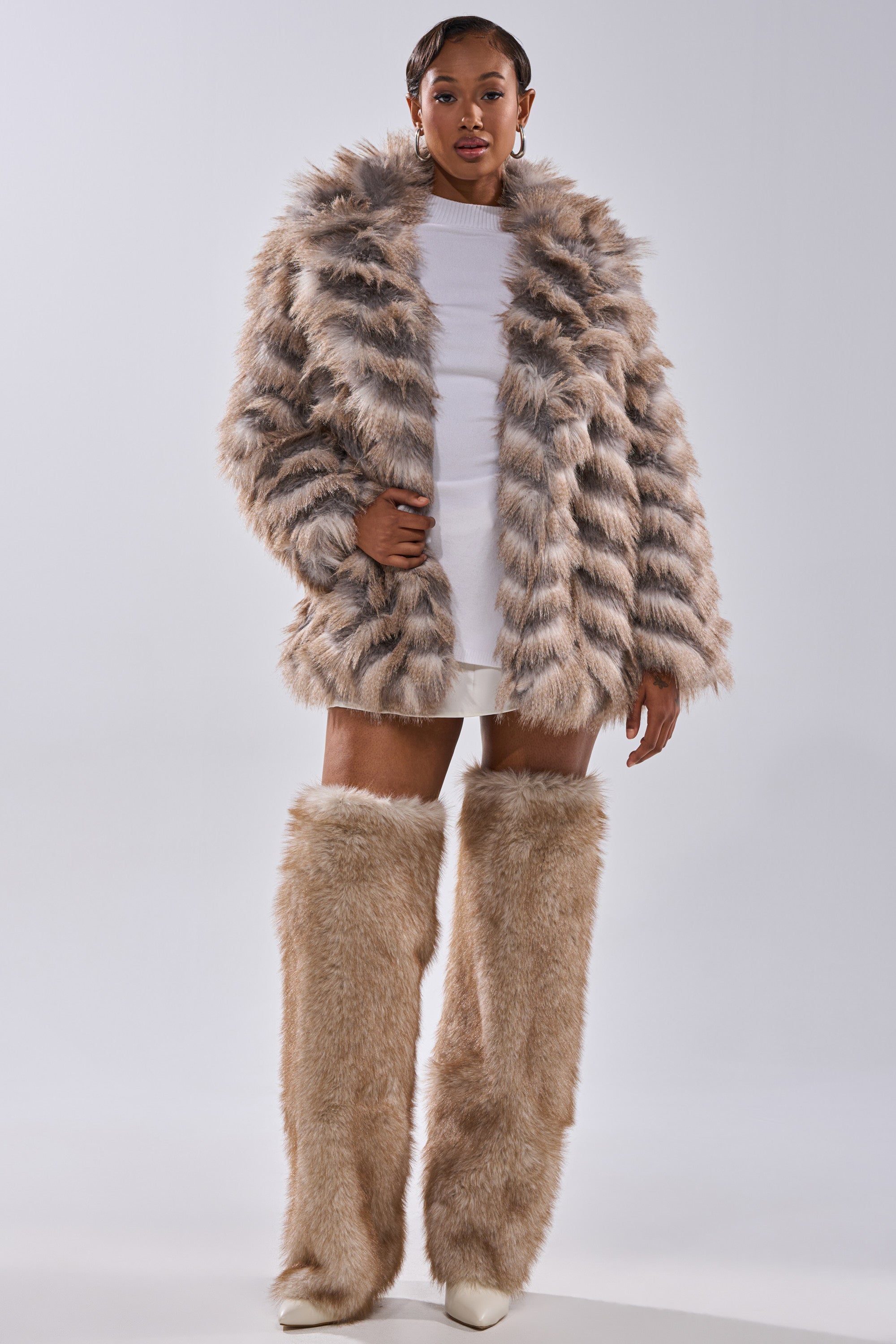 A woman faces forward in the "SHOWBIZ BABY FAUX FUR" chevron print coat and matching tall faux fur leg warmers over a white mini dress and shoes, set against a plain light background.