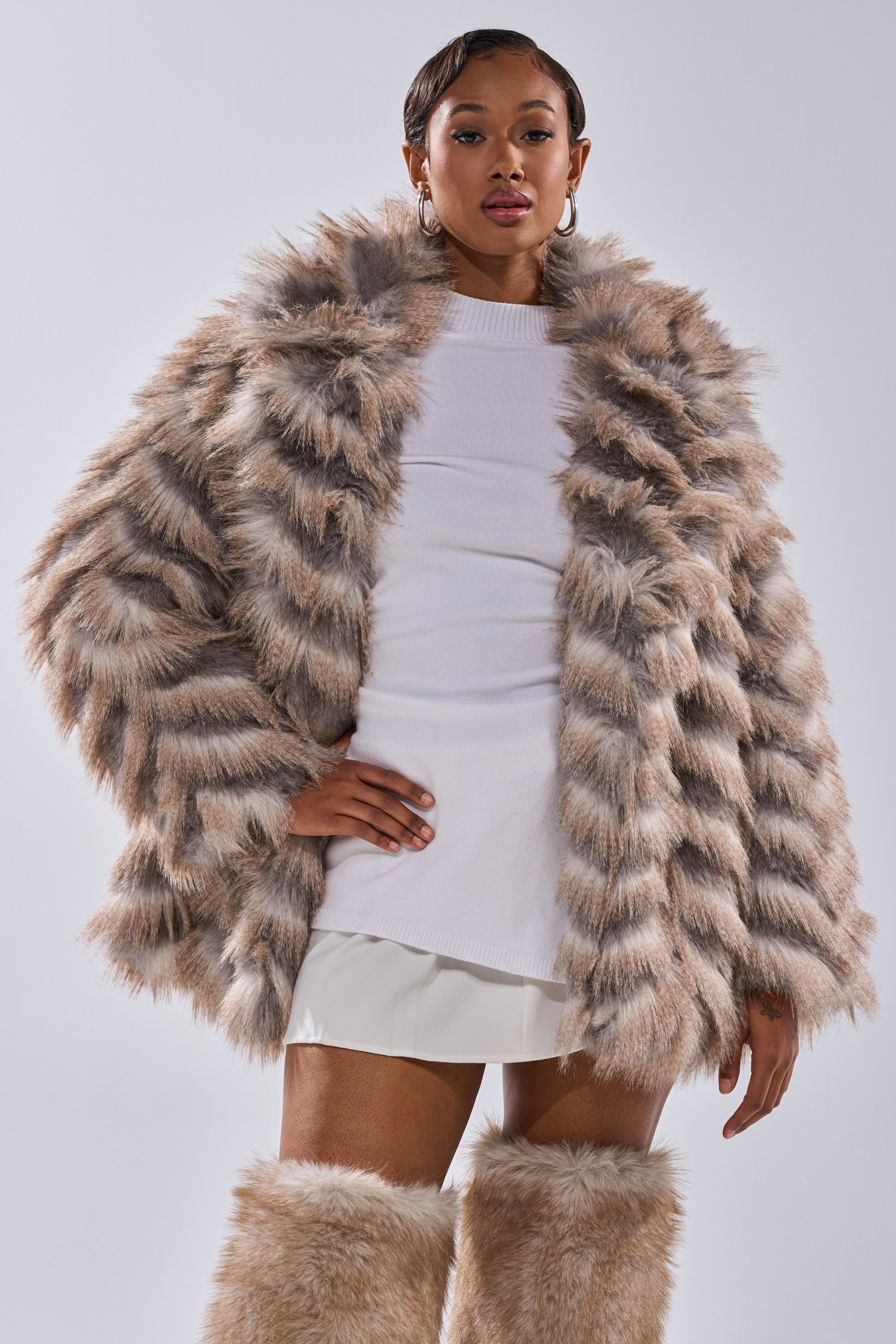 A woman confidently models the SHOWBIZ BABY FAUX FUR, a fluffy chevron print coat with an oversized collar, paired with a white mini skirt and matching fur leg warmers, posing against a plain light background.