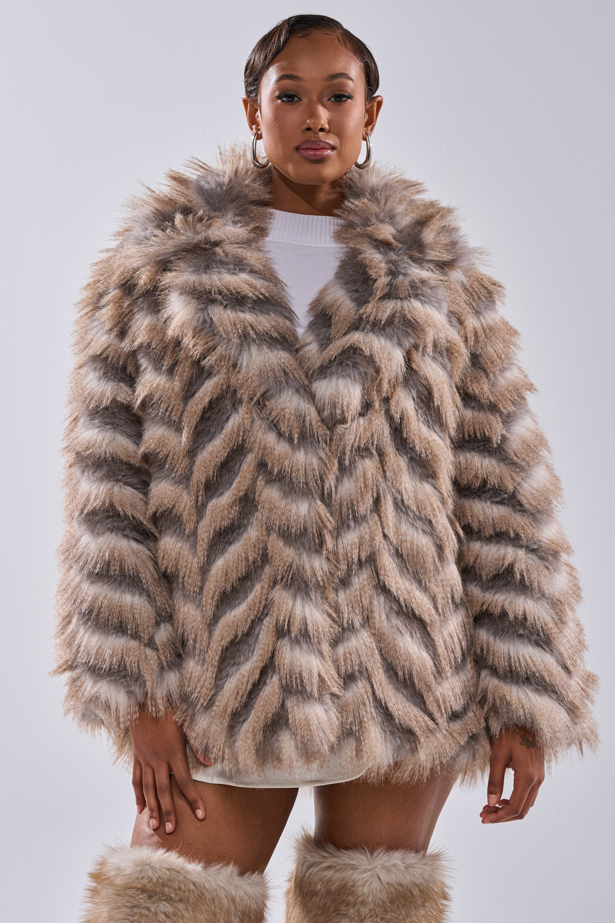 A woman with short hair and hoop earrings stands against a plain background wearing the SHOWBIZ BABY FAUX FUR coat—thick, chevron print in tan and gray—with matching fluffy boots.