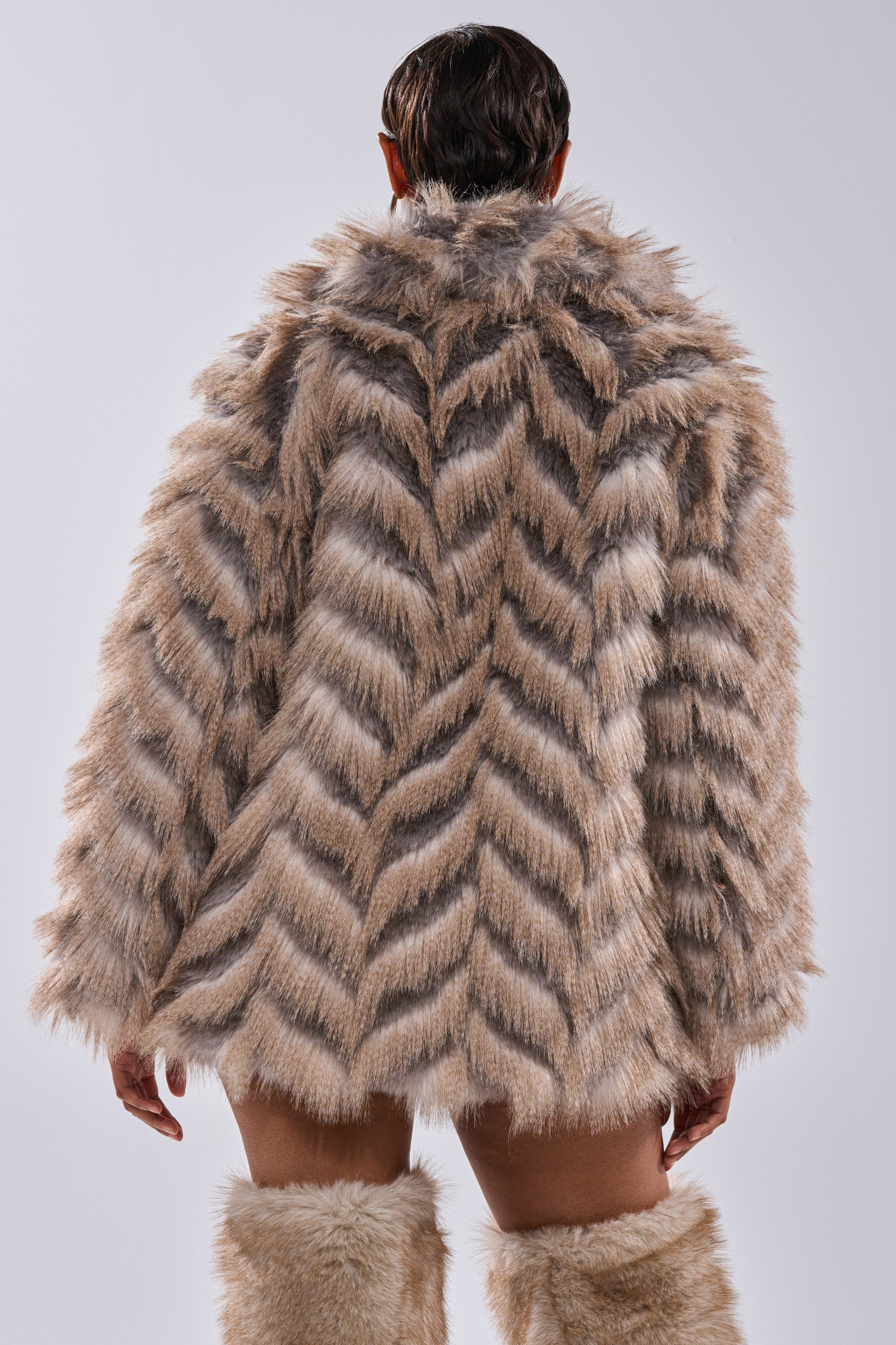 A person faces away from the camera, showcasing the SHOWBIZ BABY FAUX FUR coat in textured light brown with a longline silhouette and matching fur boots, set against a plain light background.