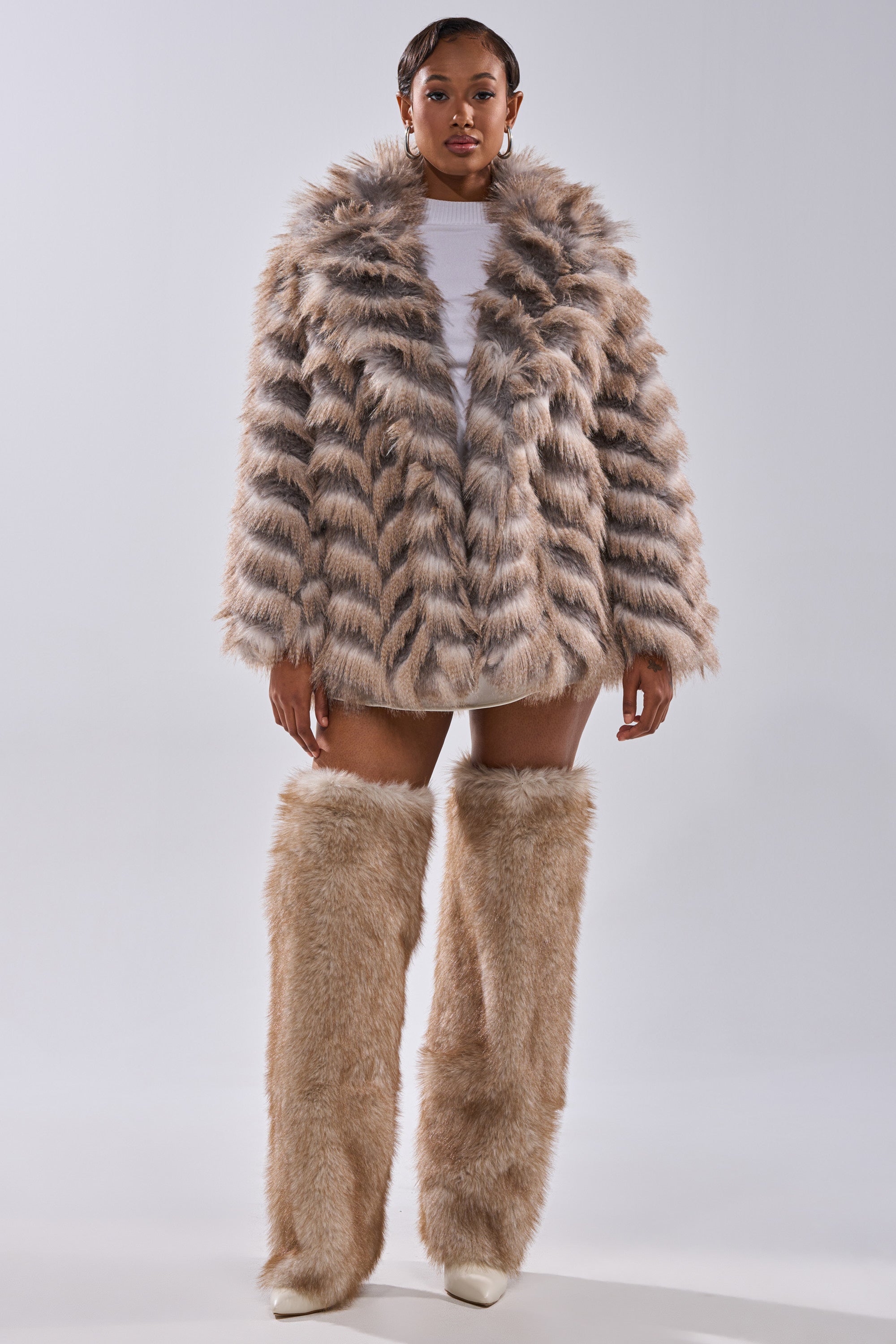 A woman faces forward in the SHOWBIZ BABY FAUX FUR coat with matching chevron faux fur leg warmers, paired with a white top and shoes, set against a plain light background.