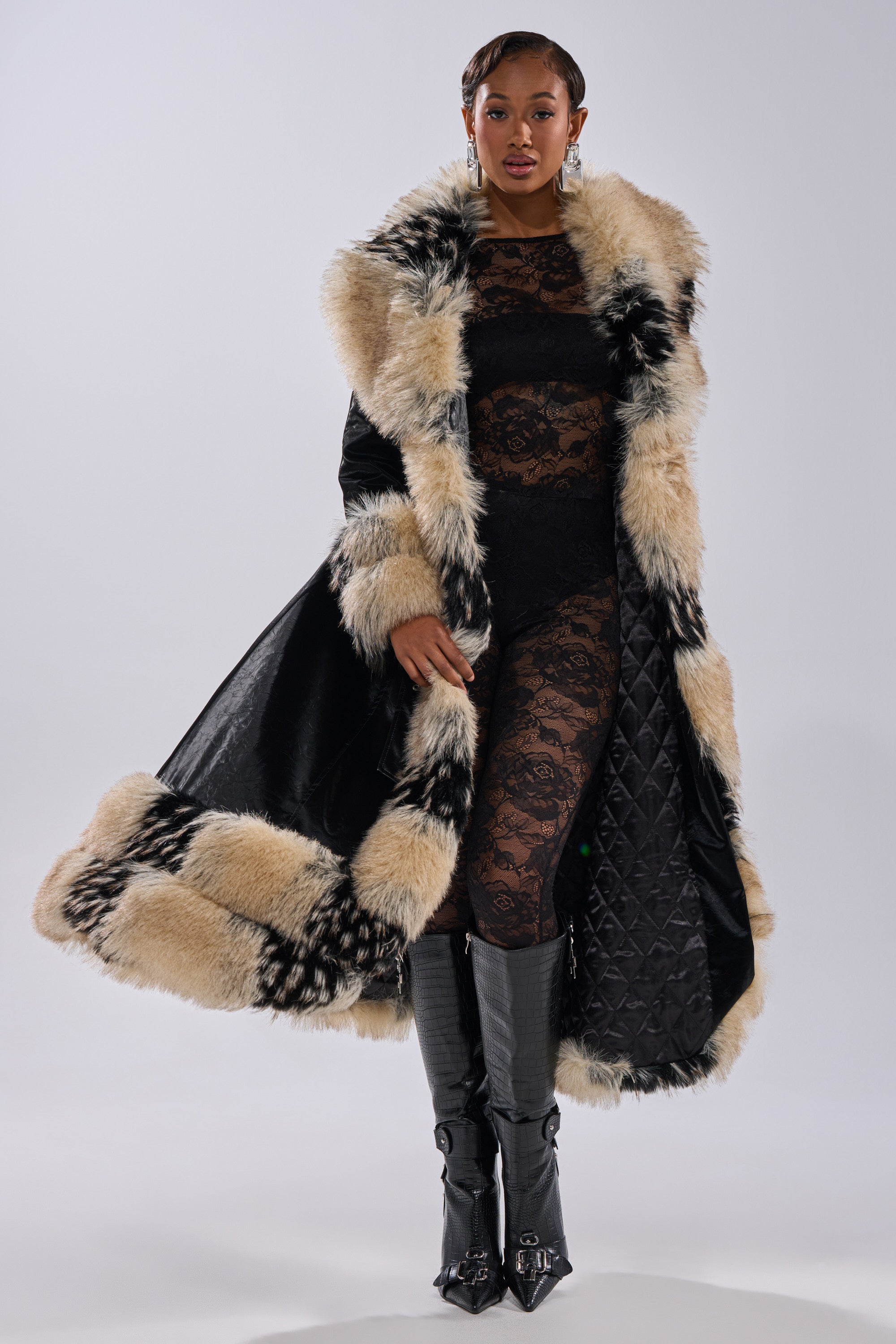A woman stands confidently in the VIVIAN FUR TRIM TRENCH IN LIQUID VELVET, featuring bold beige and black fur trim, over a sheer black lace bodysuit, paired with knee-high buckled boots and statement earrings against a neutral background.