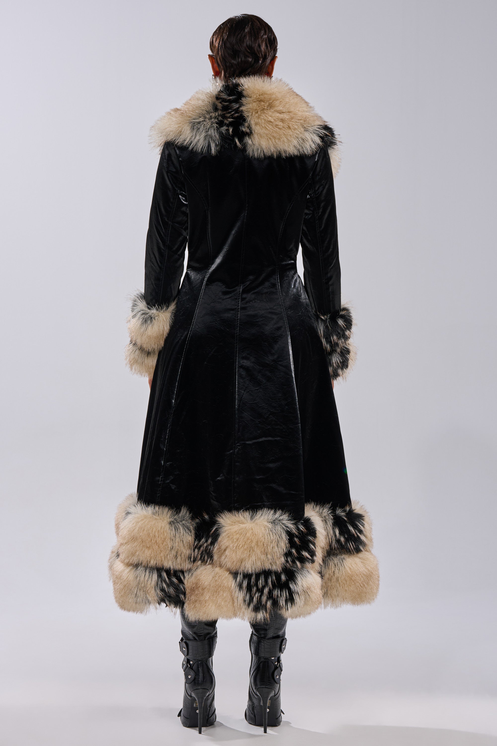 A person faces away, wearing the VIVIAN FUR TRIM TRENCH IN LIQUID VELVET with plush light faux fur trim on the collar, cuffs, and hem, paired with black knee-high boots against a plain background.