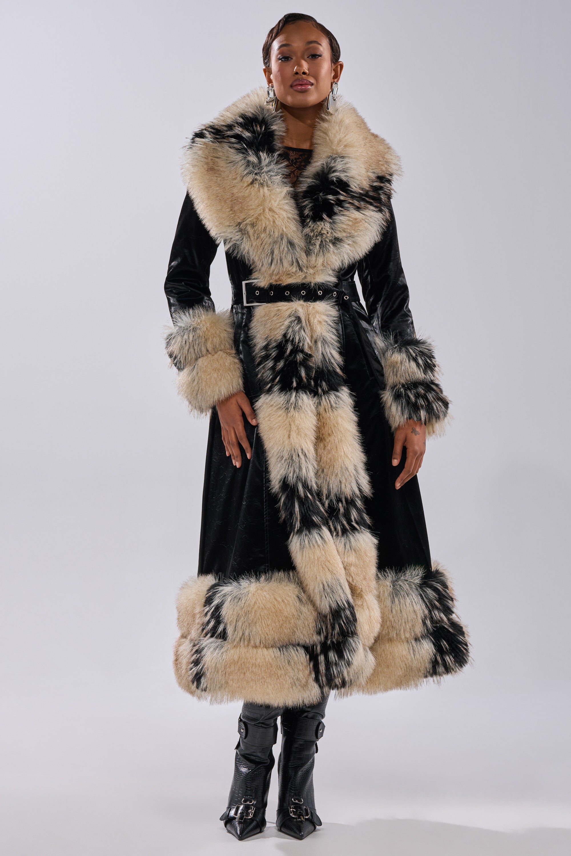 A woman faces forward wearing the VIVIAN FUR TRIM TRENCH IN LIQUID VELVET, featuring large cream and black fur accents on the collar, cuffs, and hem, styled with black boots against a plain light background.