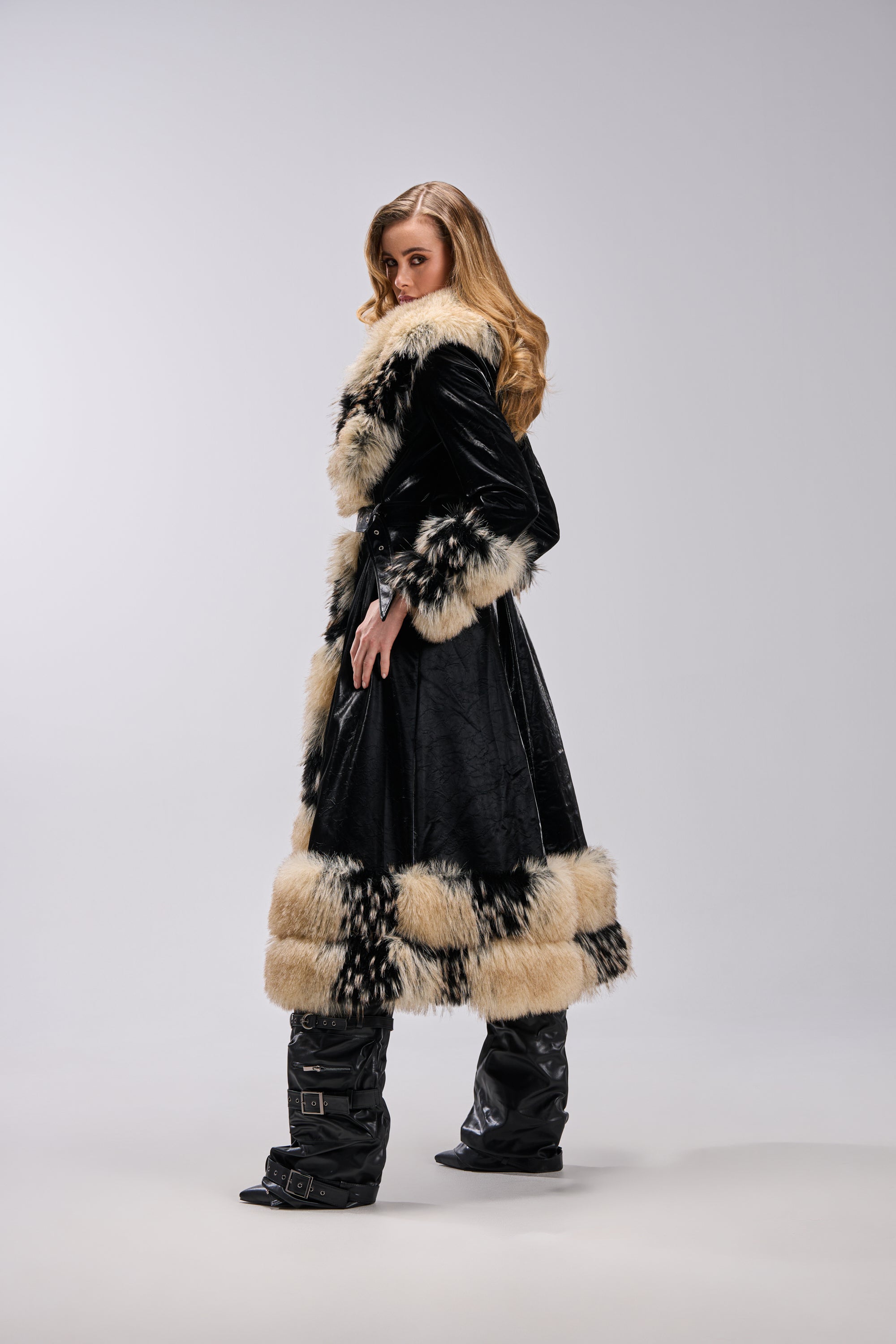 A woman with wavy blonde hair models the VIVIAN FUR TRIM TRENCH IN LIQUID VELVET, featuring black liquid velvet and beige-black fur trim, paired with shiny black boots against a neutral backdrop.