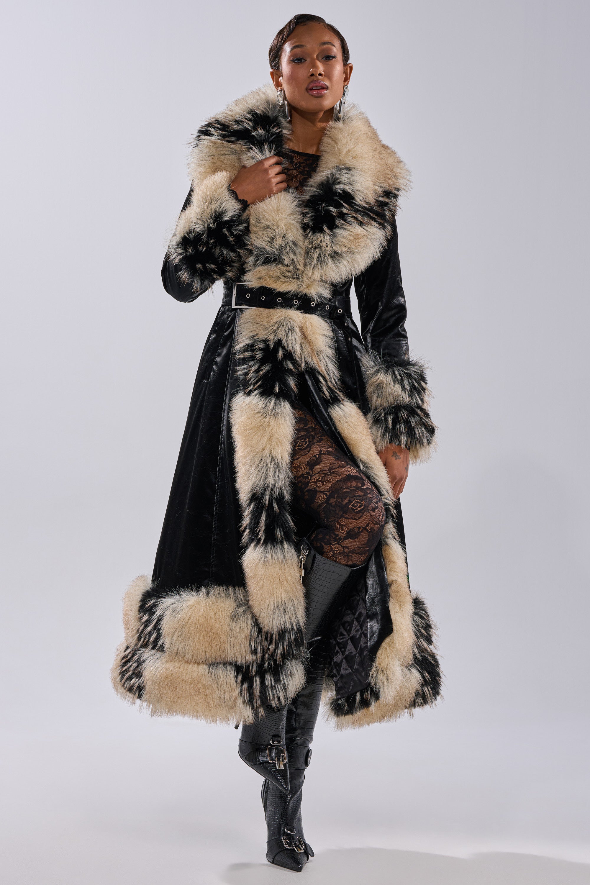 A woman poses confidently in the VIVIAN FUR TRIM TRENCH IN LIQUID VELVET, its bold cream and black faux fur trim adding drama. She pairs it with knee-high boots and a lace dress against a plain light gray background.