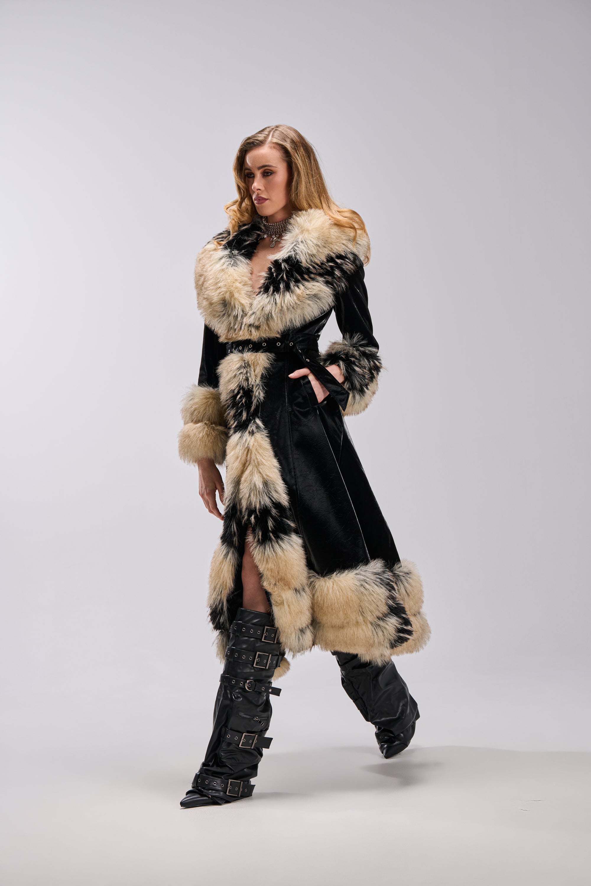 A woman confidently walks against a light gray background, wearing the VIVIAN FUR TRIM TRENCH IN LIQUID VELVET paired with black knee-high buckled boots.