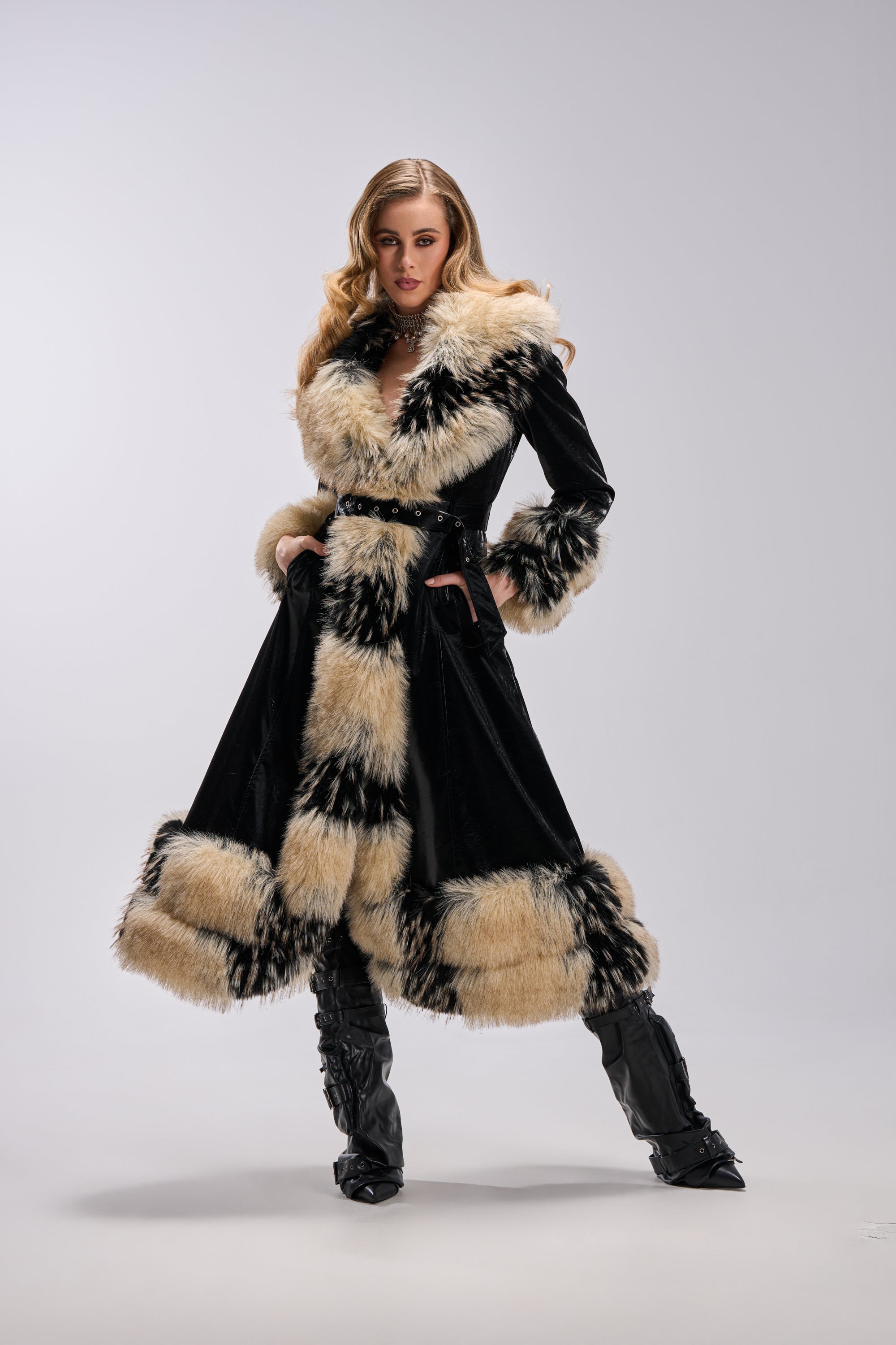 A woman stands confidently in the VIVIAN FUR TRIM TRENCH IN LIQUID VELVET, featuring dramatic tan and black fur accents and matching cuffs, paired with tall black boots against a plain light grey background.