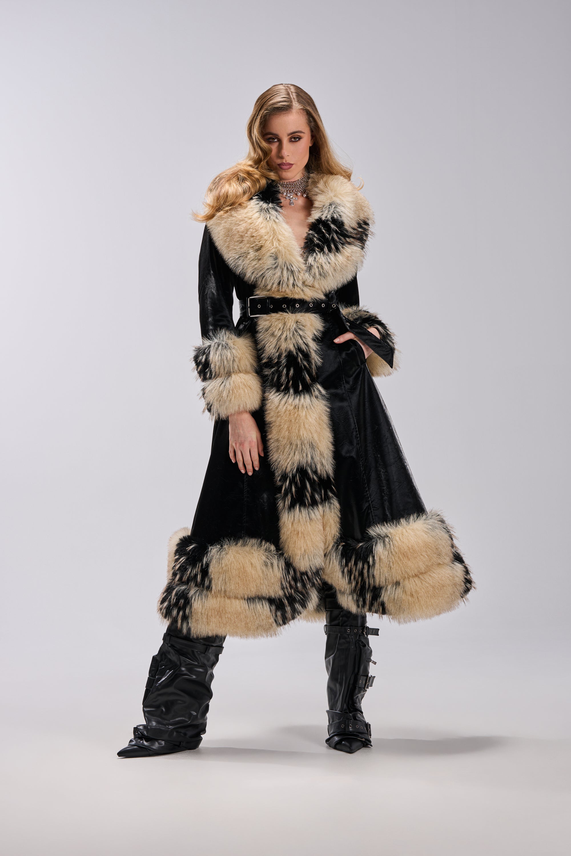 A woman models the VIVIAN FUR TRIM TRENCH IN LIQUID VELVET, featuring bold cream and black fur accents on the collar, cuffs, and hem. She pairs it with black boots, her wavy hair contrasting against a plain light background.