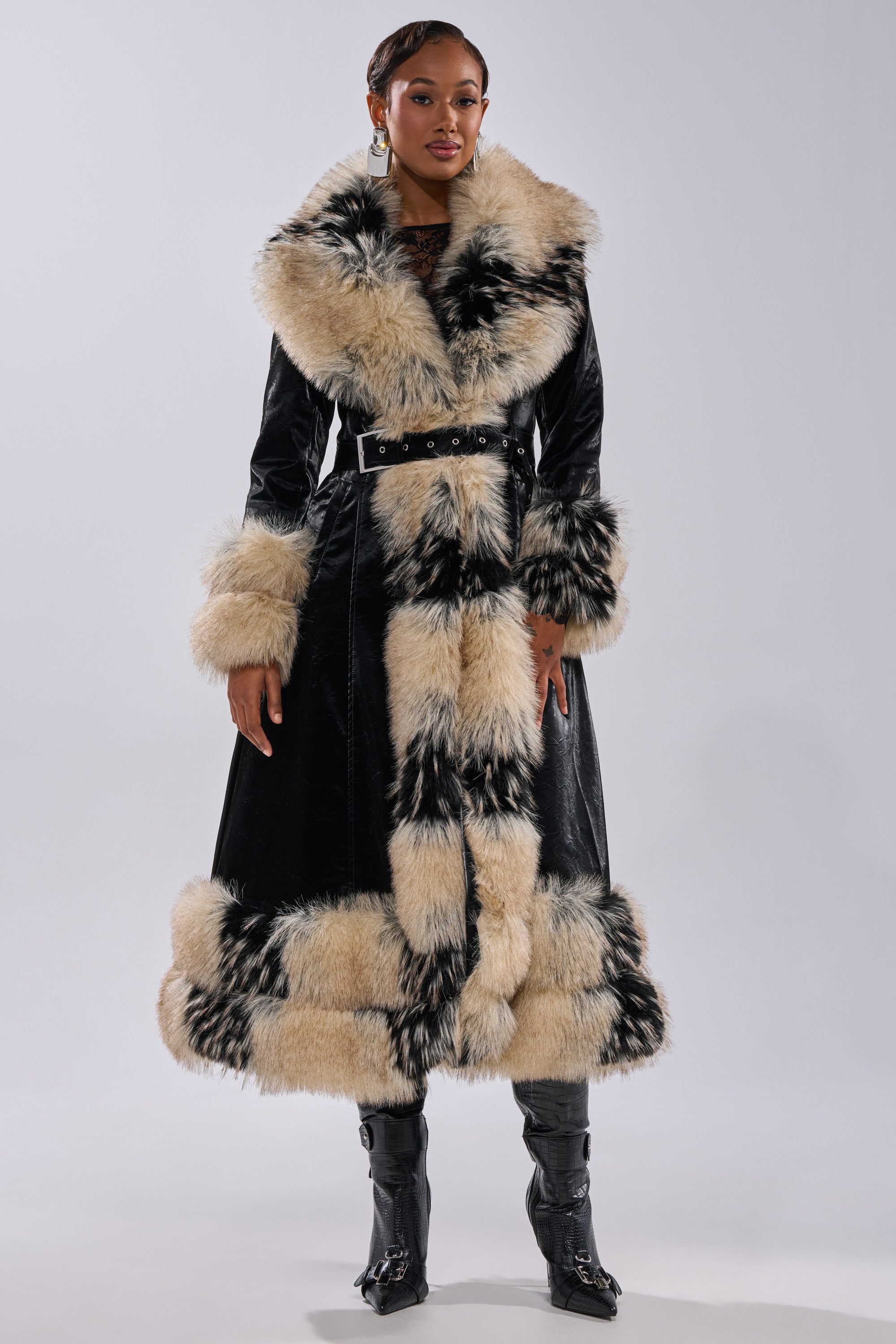 A woman models the VIVIAN FUR TRIM TRENCH IN LIQUID VELVET, featuring bold beige and black fur at the collar, cuffs, and hem. She accessorizes with earrings and black boots, posing against a plain light background.
