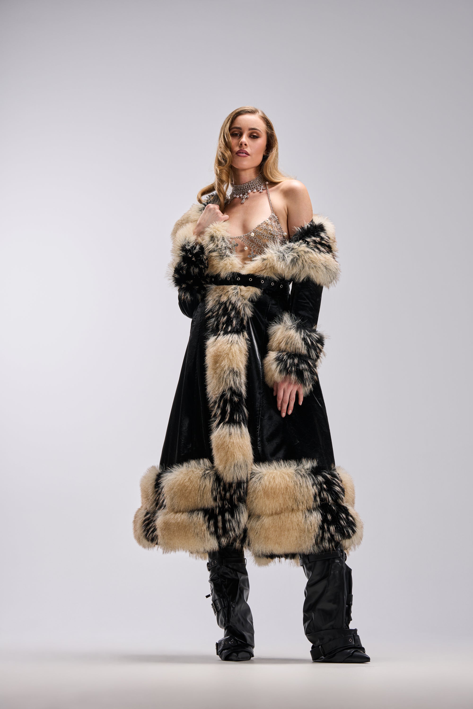 A woman poses confidently in the VIVIAN FUR TRIM TRENCH IN LIQUID VELVET, paired with a sparkling top, high boots, and statement jewelry against a plain light background.