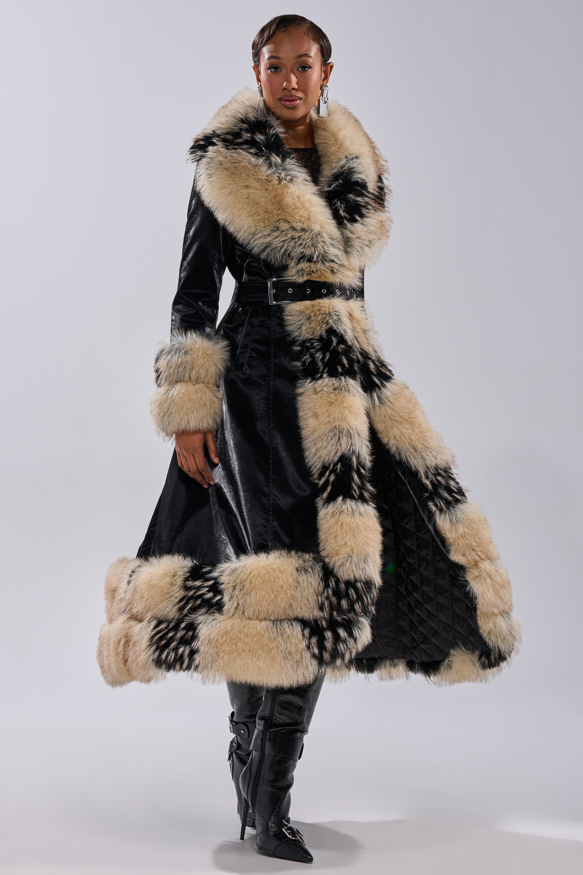 A woman stands against a plain light background wearing the VIVIAN FUR TRIM TRENCH IN LIQUID VELVET, featuring thick cream and black fur trim at the collar, cuffs, and hem, belted at the waist, paired with tall black boots.
