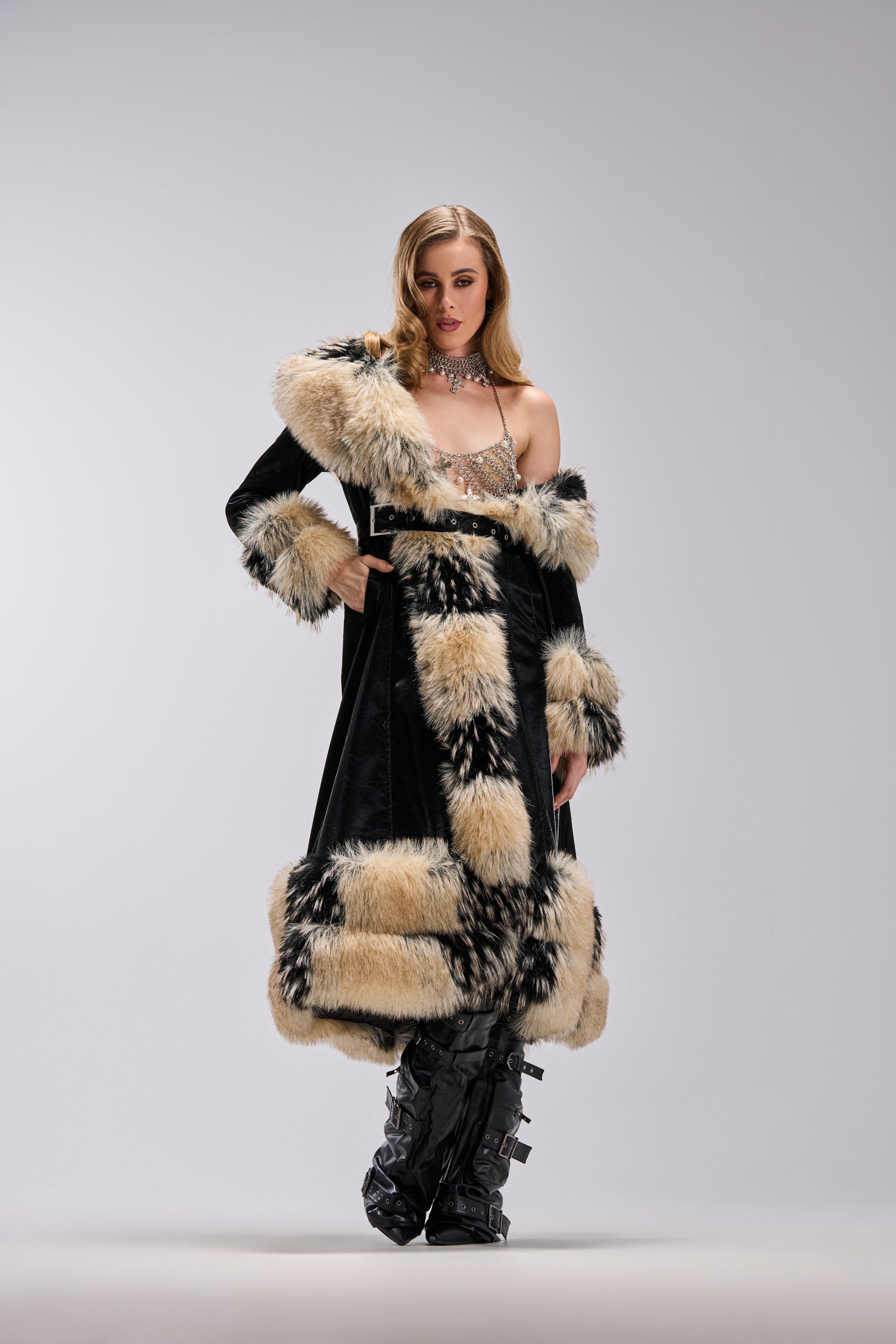 A woman stands confidently in the VIVIAN FUR TRIM TRENCH IN LIQUID VELVET, featuring dramatic tan and black fur accents, a sparkling top, and knee-high black buckle boots against a plain light gray background.