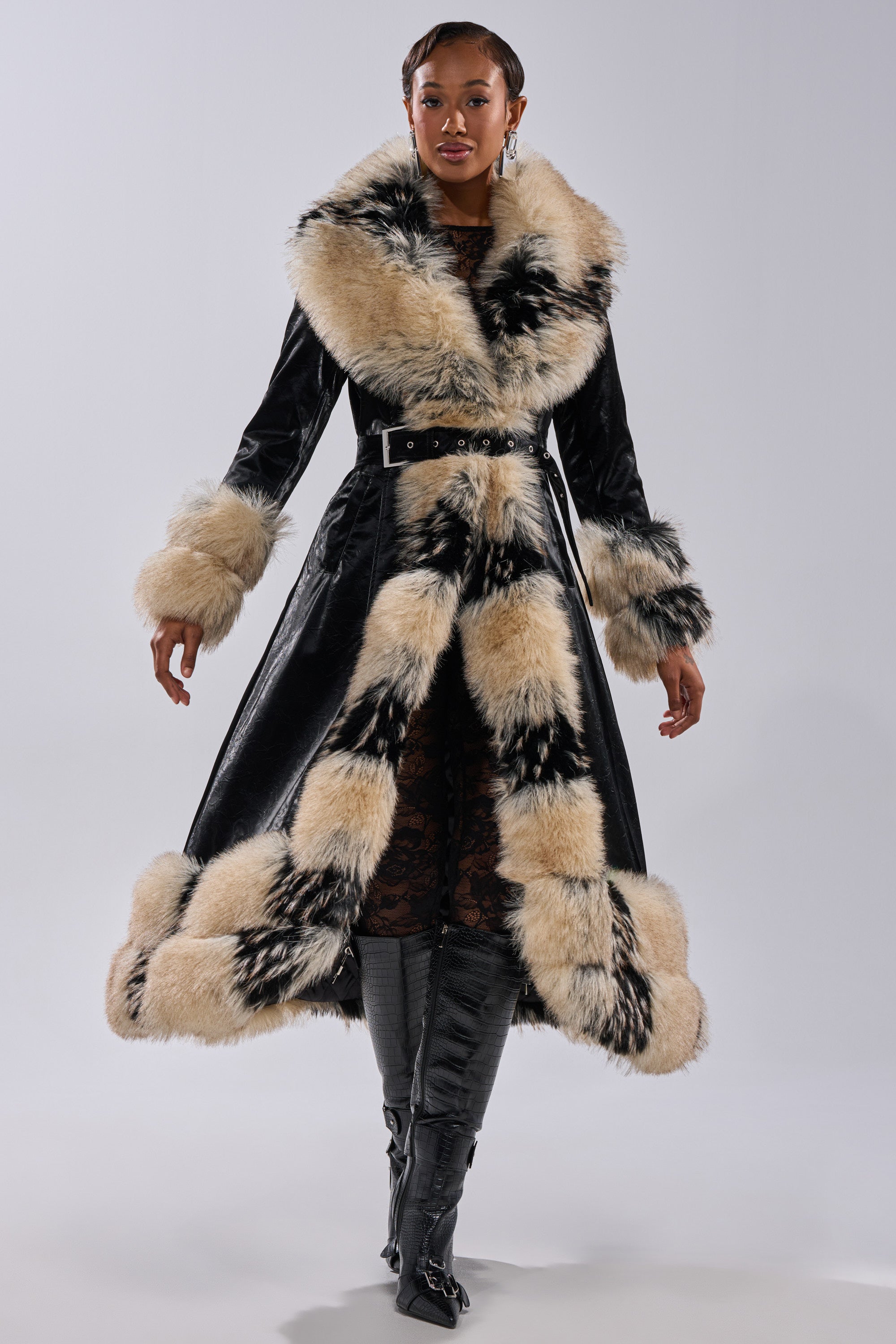 A woman poses confidently in the VIVIAN FUR TRIM TRENCH IN LIQUID VELVET, featuring beige and black faux fur on the collar, cuffs, and hem, belted at the waist, styled with knee-high black boots against a plain background.