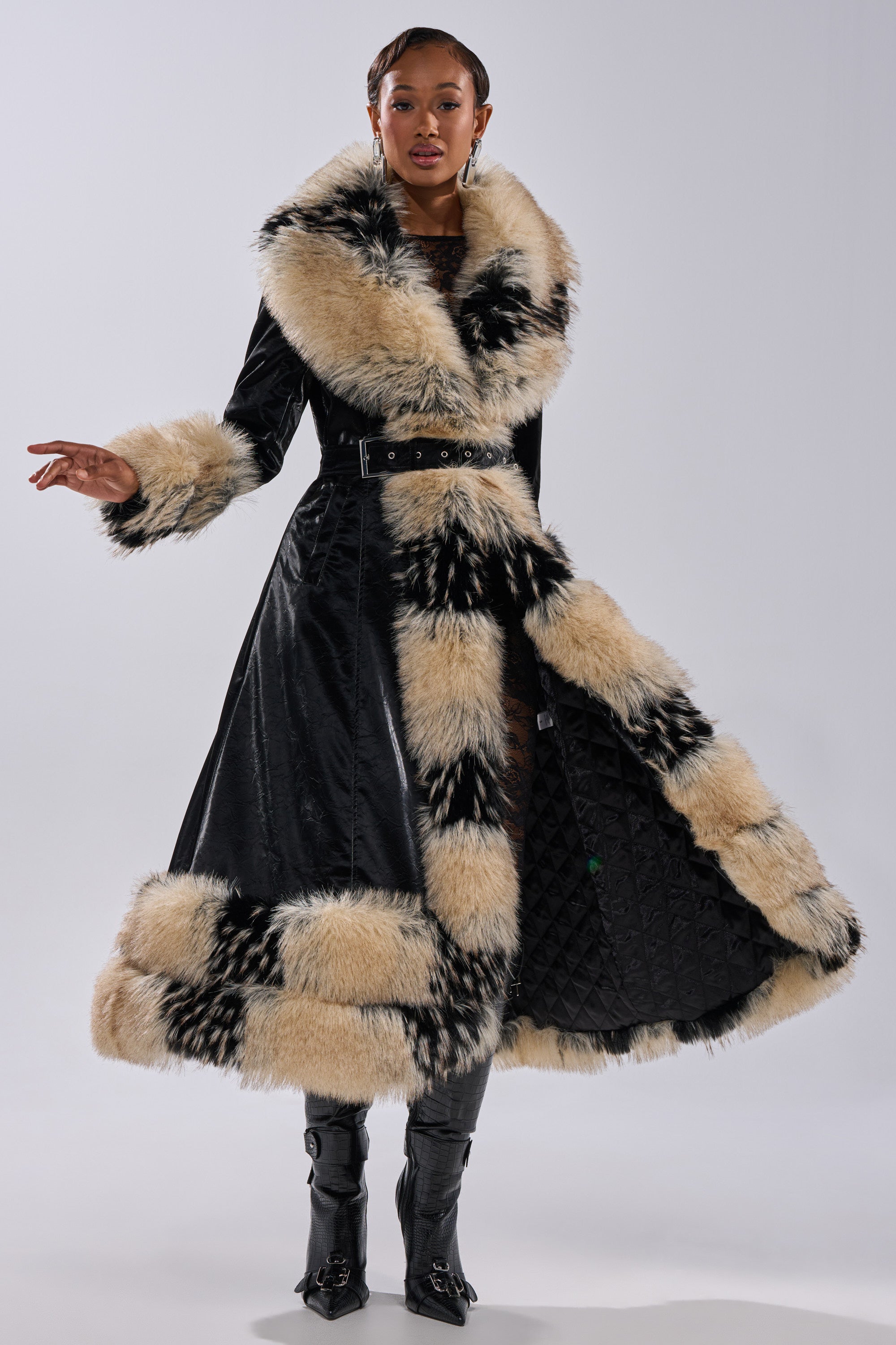 A person stands confidently in the VIVIAN FUR TRIM TRENCH IN LIQUID VELVET, a long black coat with wide cream fur trim, belted waist, tall black boots, and an open front revealing a quilted lining.