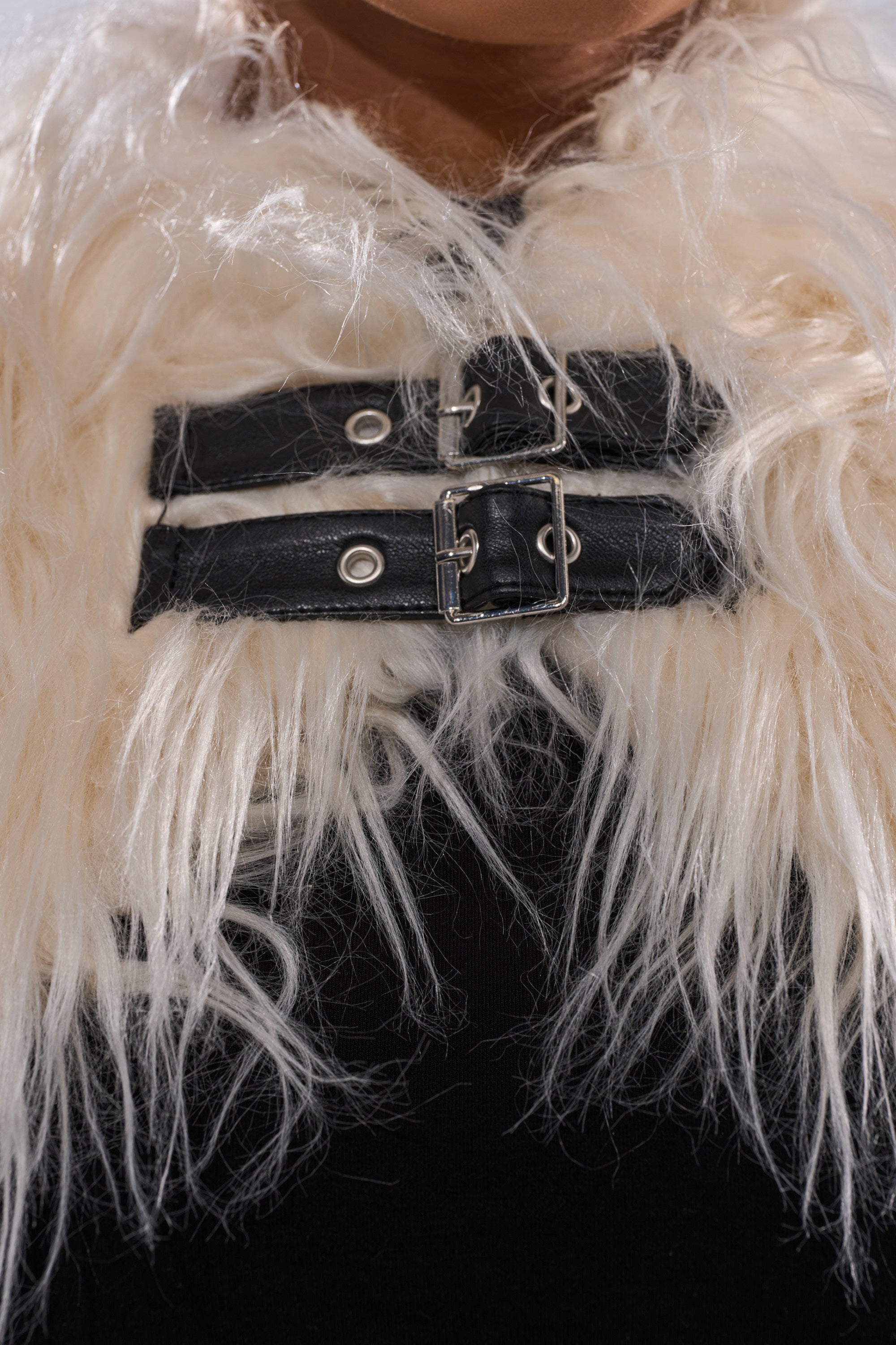 Close-up of a person wearing the PHIA FAUX FUR BOLERO, a creamy cropped jacket with two black leather buckle straps and silver hardware across the chest, layered over a black top for a bold statement look.
