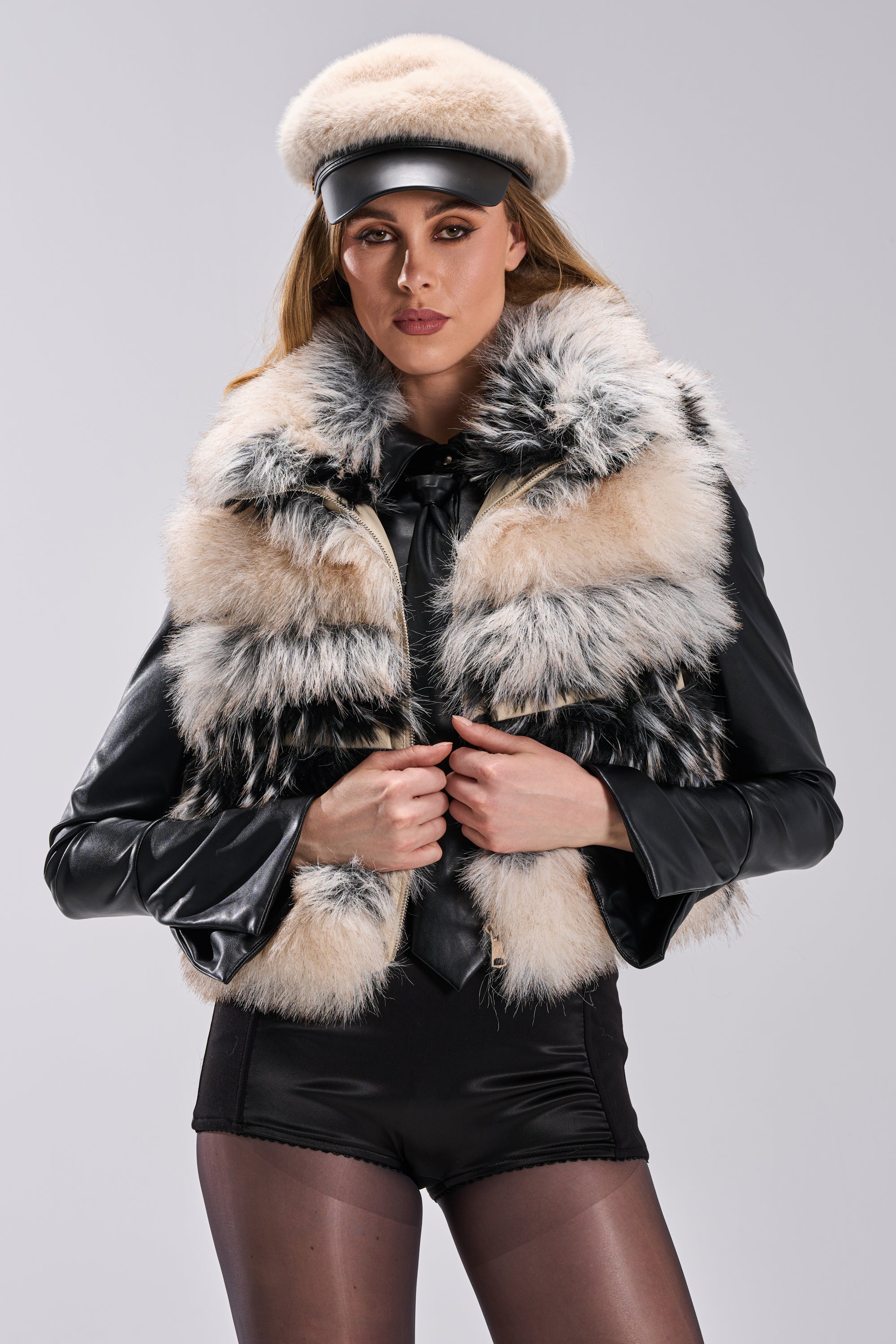 Wearing the KIKI FAUX FUR VEST with an oversized collar over a black leather jacket, paired with black shorts, sheer tights, and a cream fur beret with a black visor, a woman poses confidently against a light gray background.