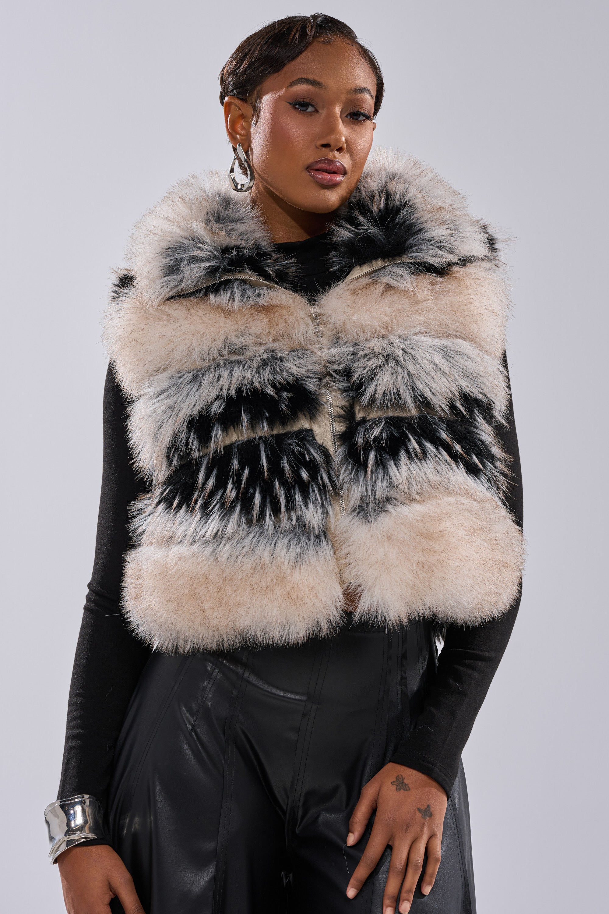 A woman models the KIKI FAUX FUR VEST, featuring a striped design and oversized collar, over a black long-sleeve top and pants. She wears earrings, a bracelet, and has short, styled hair, all set against a plain light background.