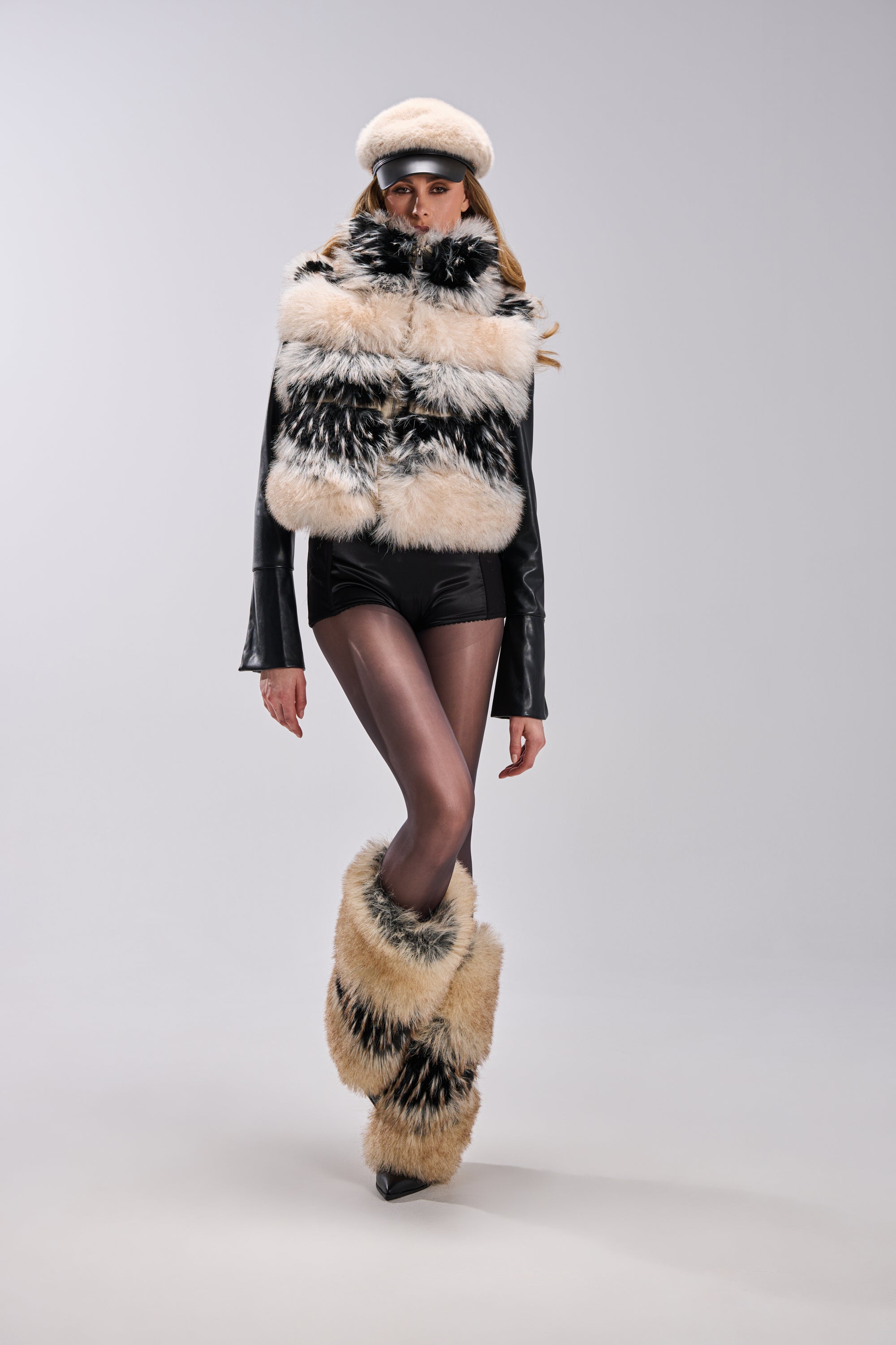 A model in the KIKI FAUX FUR VEST with a matching faux fur hat, black leather shorts, sheer black tights, and coordinating faux fur leg warmers with bungee cord walks against a plain light gray background.