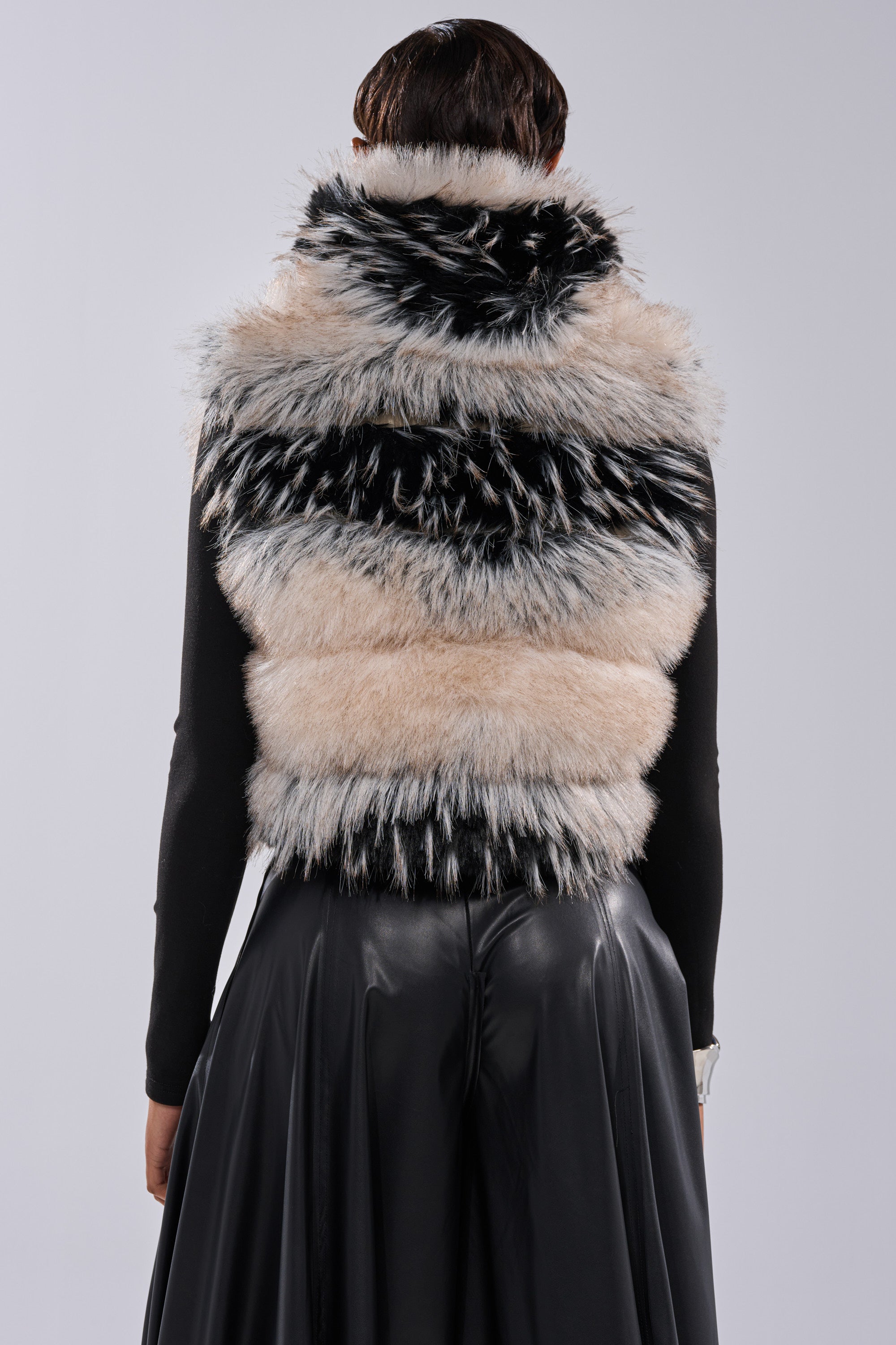 A person shown from the back against a plain light background wears a black long-sleeve top, black skirt, and the KIKI FAUX FUR VEST—a striped two-tone faux fur vest featuring white, gray, and black accents.