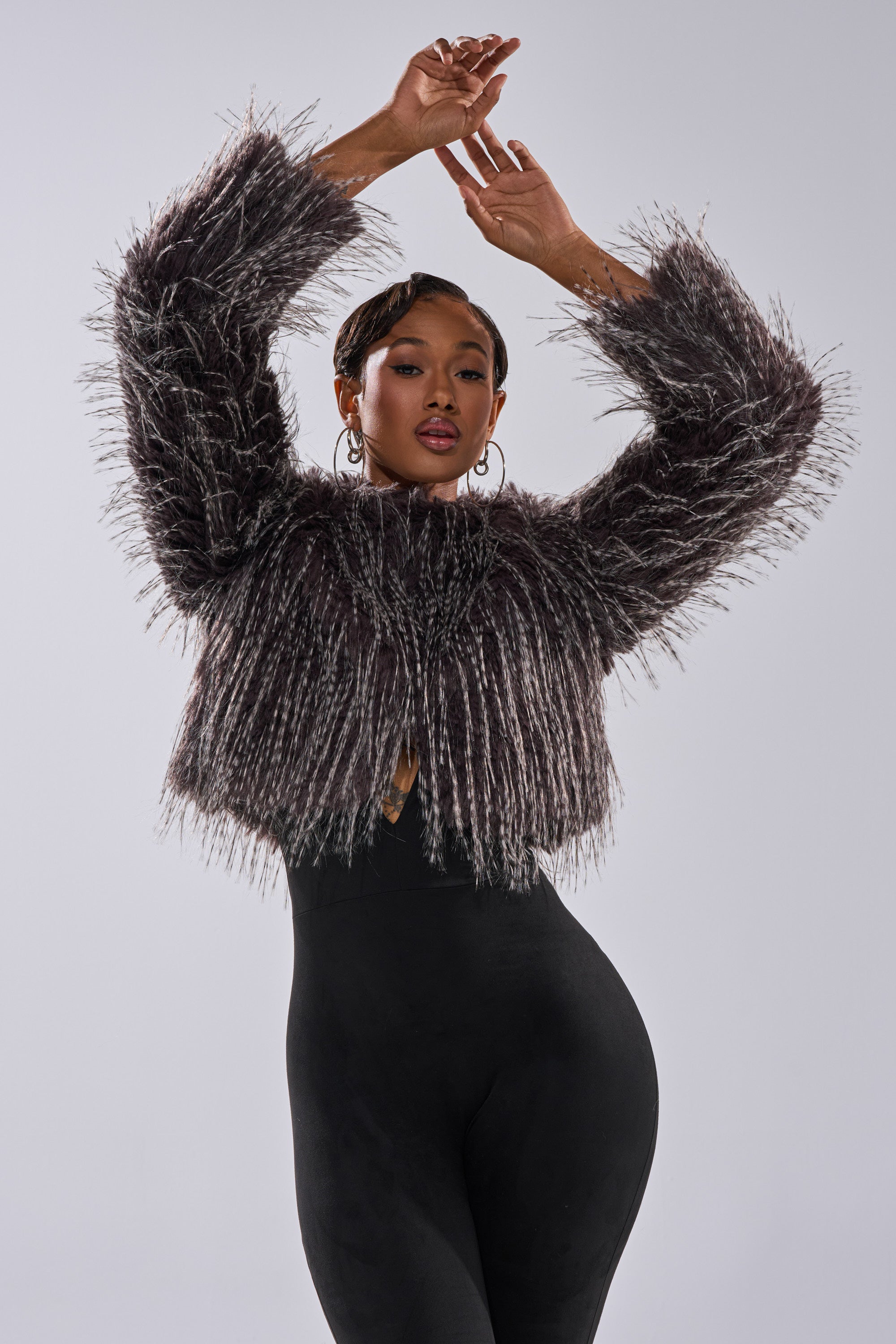 A woman poses with arms raised in the BIRDS OF A FEATHER SHAGGY FAUX FUR jacket, featuring a boxy fit, paired with black fitted pants against a plain light background.