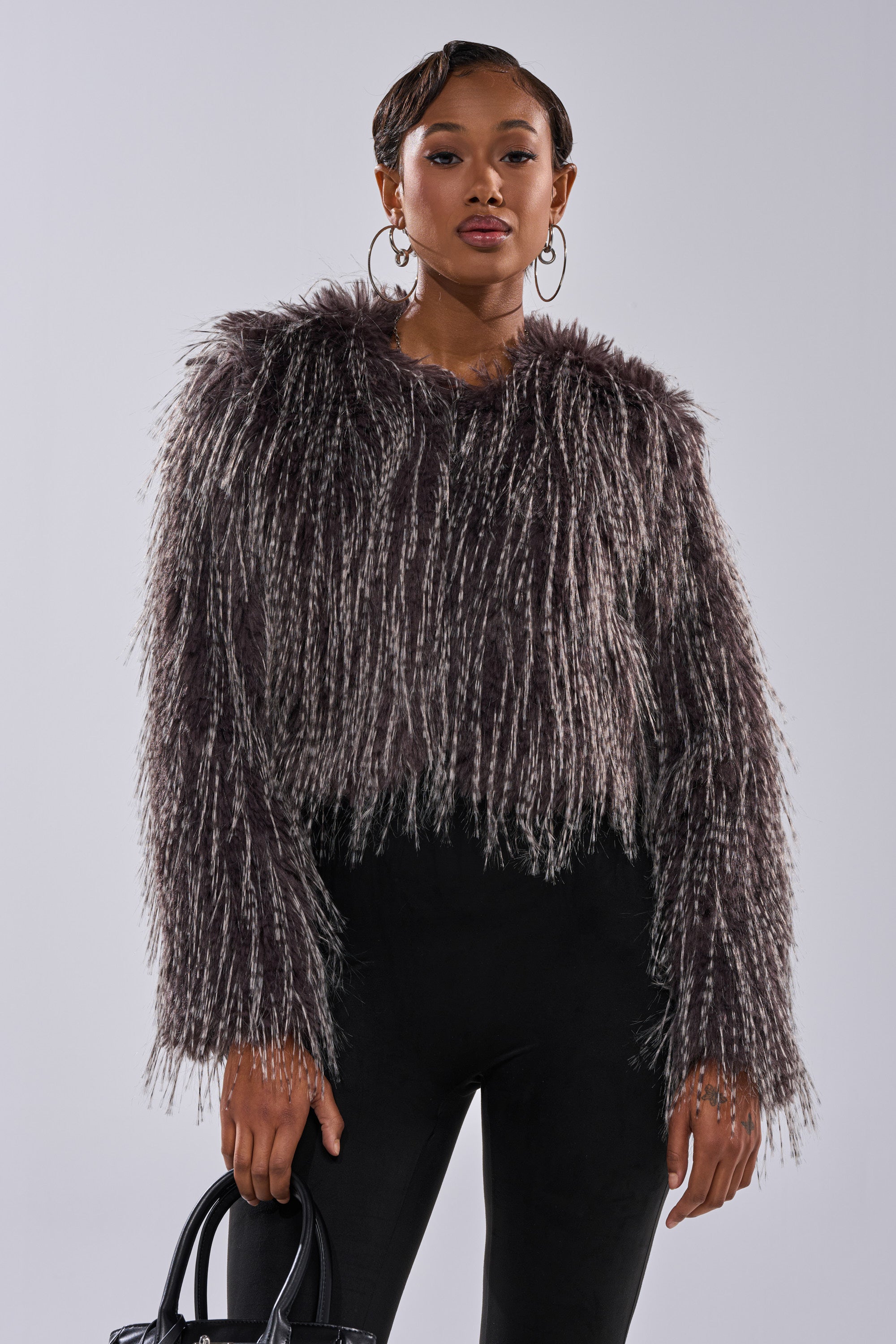A woman showcases the BIRDS OF A FEATHER SHAGGY FAUX FUR jacket in dark brown, featuring a textured, boxy fit. She pairs it with black pants and a black handbag, accessorized with hoop earrings, against a light backdrop.