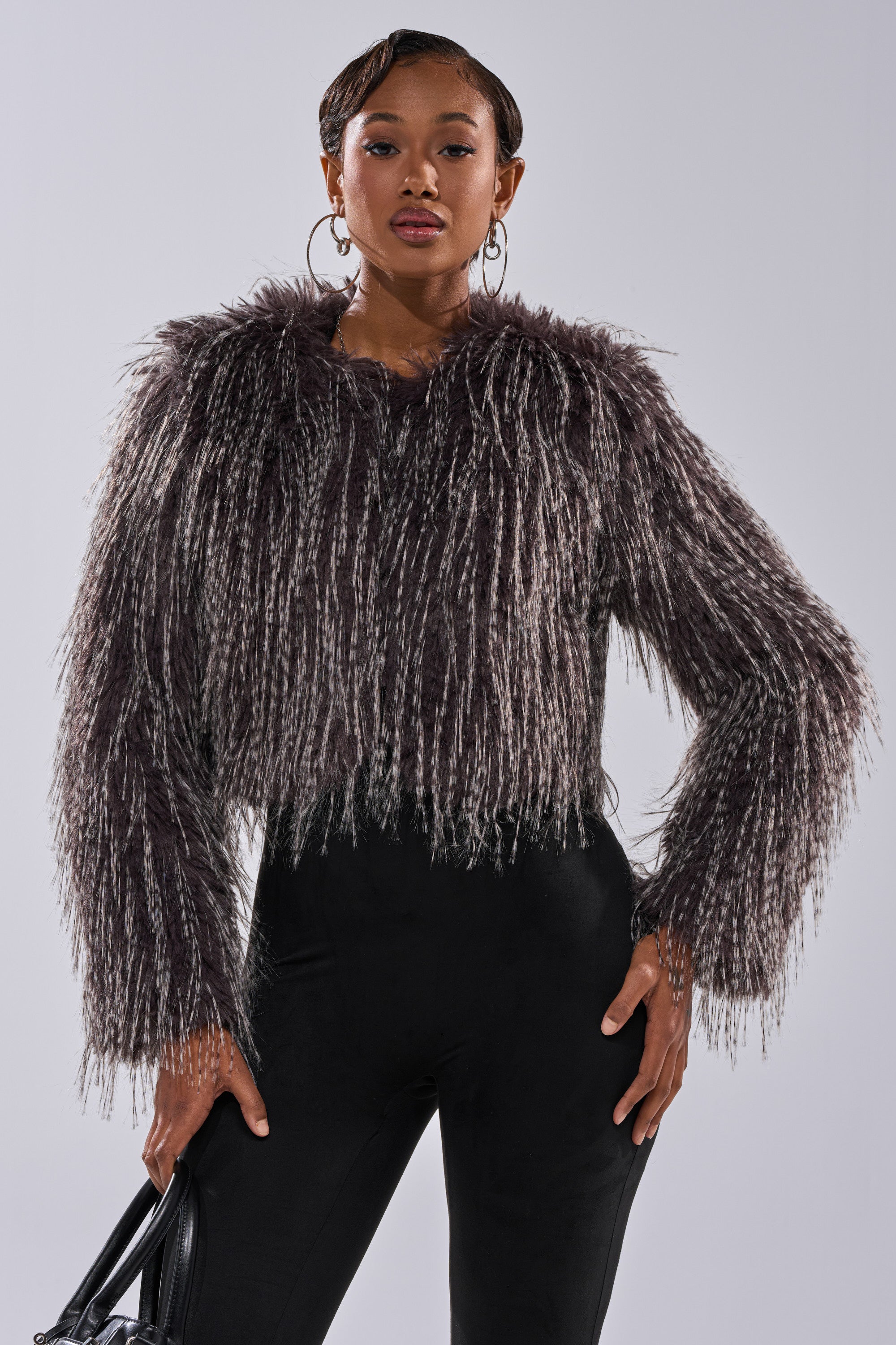 A woman poses confidently in the BIRDS OF A FEATHER SHAGGY FAUX FUR jacket with black pants and a black handbag, her short styled hair, hoop earrings, and poised expression set against a plain background.