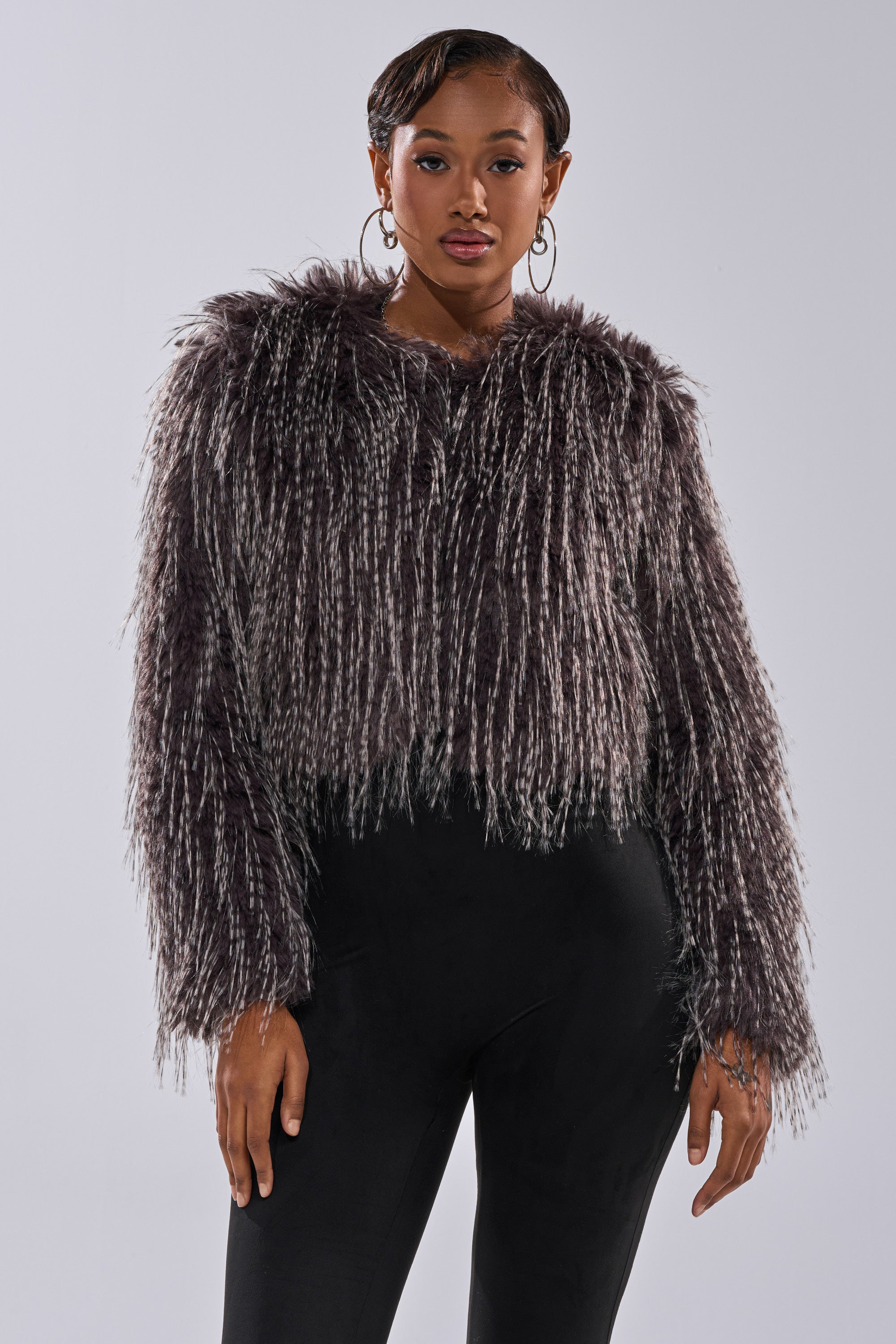 A woman faces forward, wearing the BIRDS OF A FEATHER SHAGGY FAUX FUR jacket in dark gray and black, paired with fitted black pants and hoop earrings, standing against a plain light background with a neutral expression.