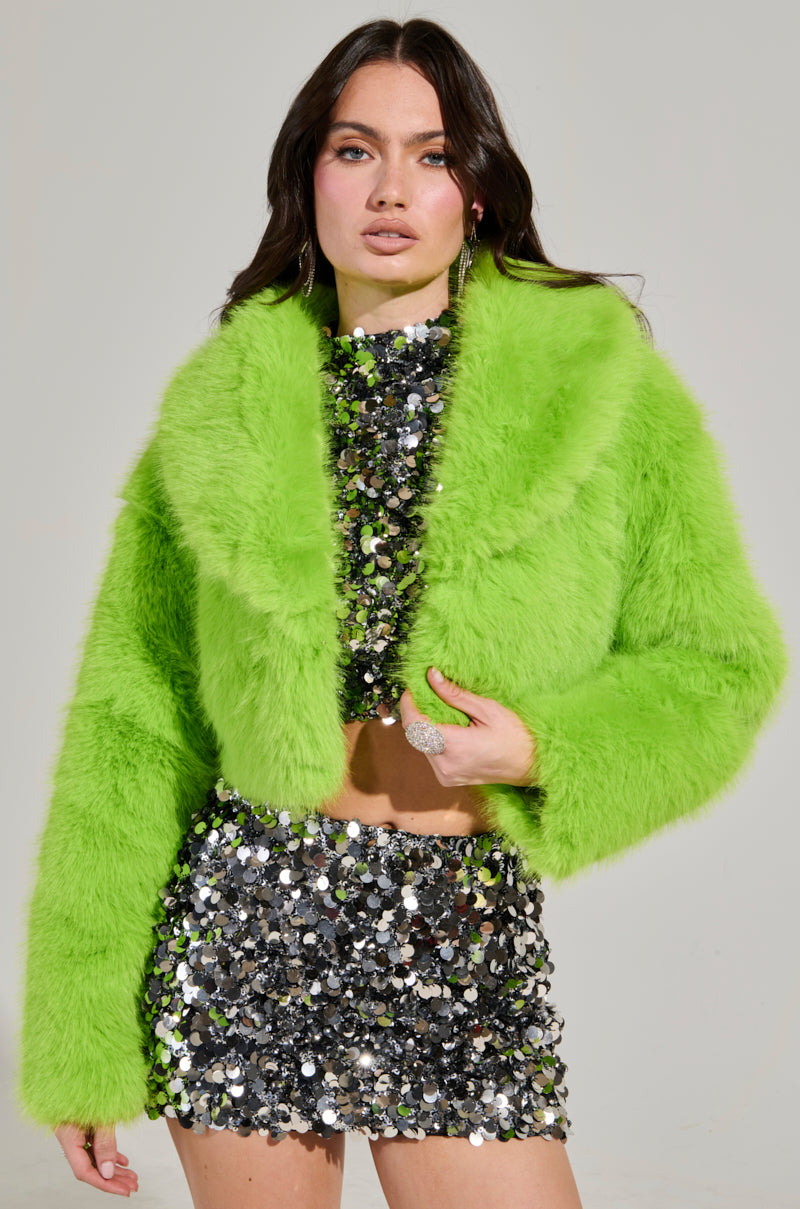 Wearing the LIGHTS OUT CROPPED FAUX FUR JACKET IN LIME, someone confidently showcases their style, layering it over a dazzling silver sequin crop top and skirt set. They pose boldly against a neutral backdrop, highlighting their fashion-forward look.