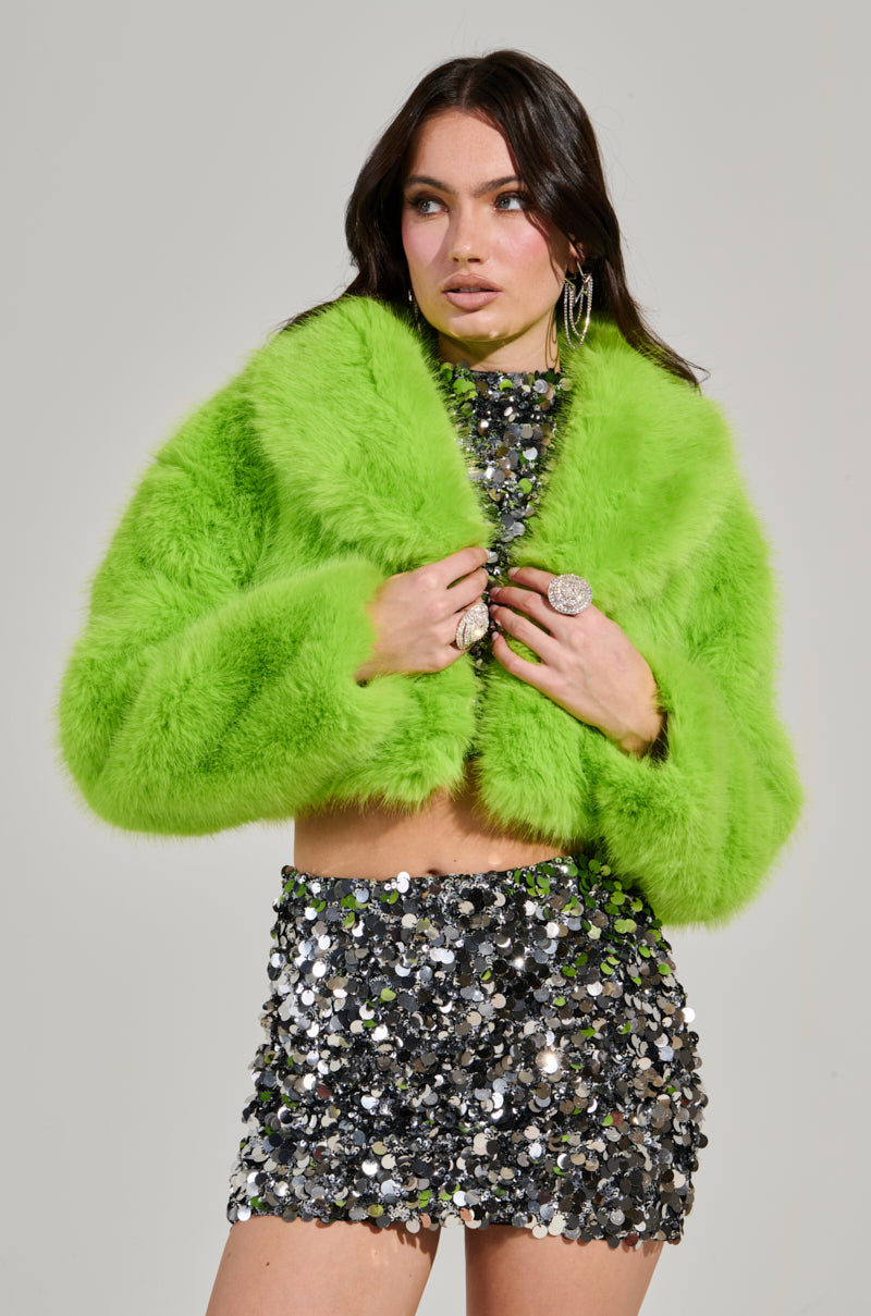 A person strikes a confident pose in the LIGHTS OUT CROPPED FAUX FUR JACKET IN LIME, featuring an oversized collar and a shiny silver sequin skirt. The plain background accentuates the bold fashion ensemble and large hoop earrings.