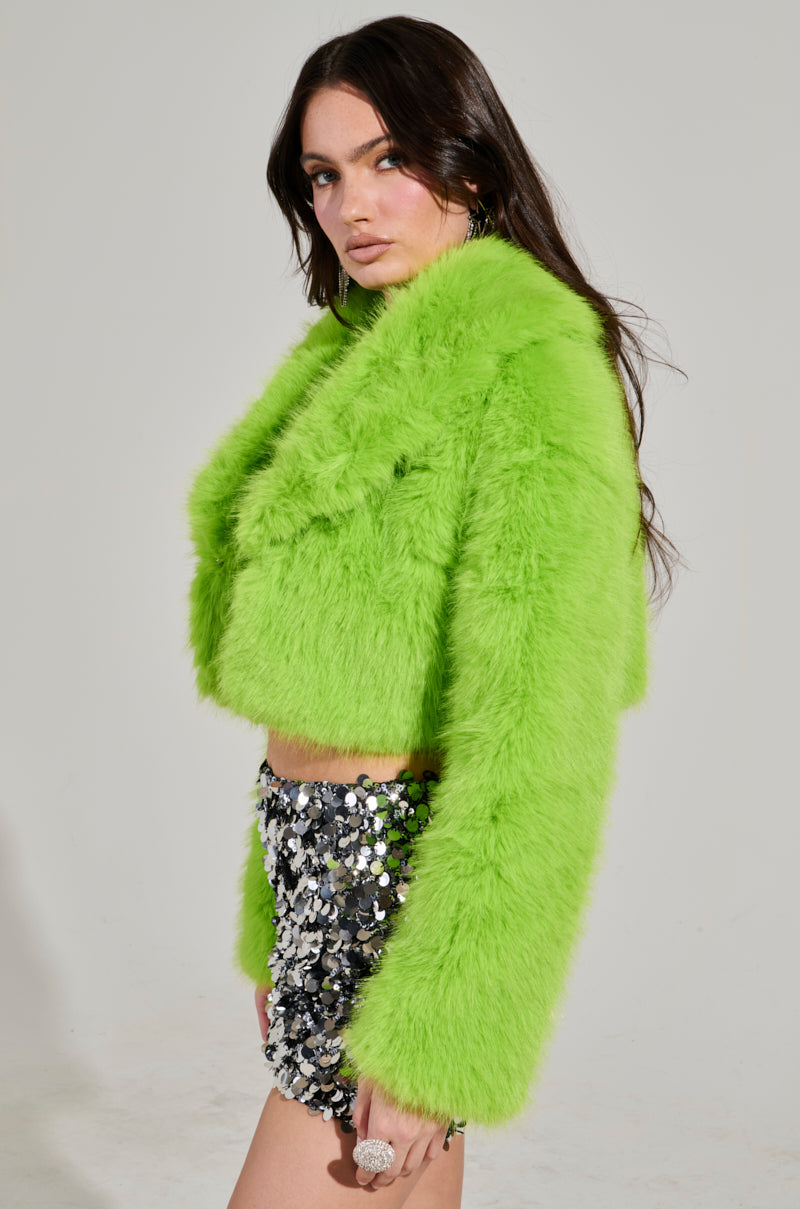 A woman showcases the LIGHTS OUT Cropped Faux Fur Jacket in Lime, paired with sequin shorts against a plain background. Her long hair cascades freely, complemented by subtle makeup.