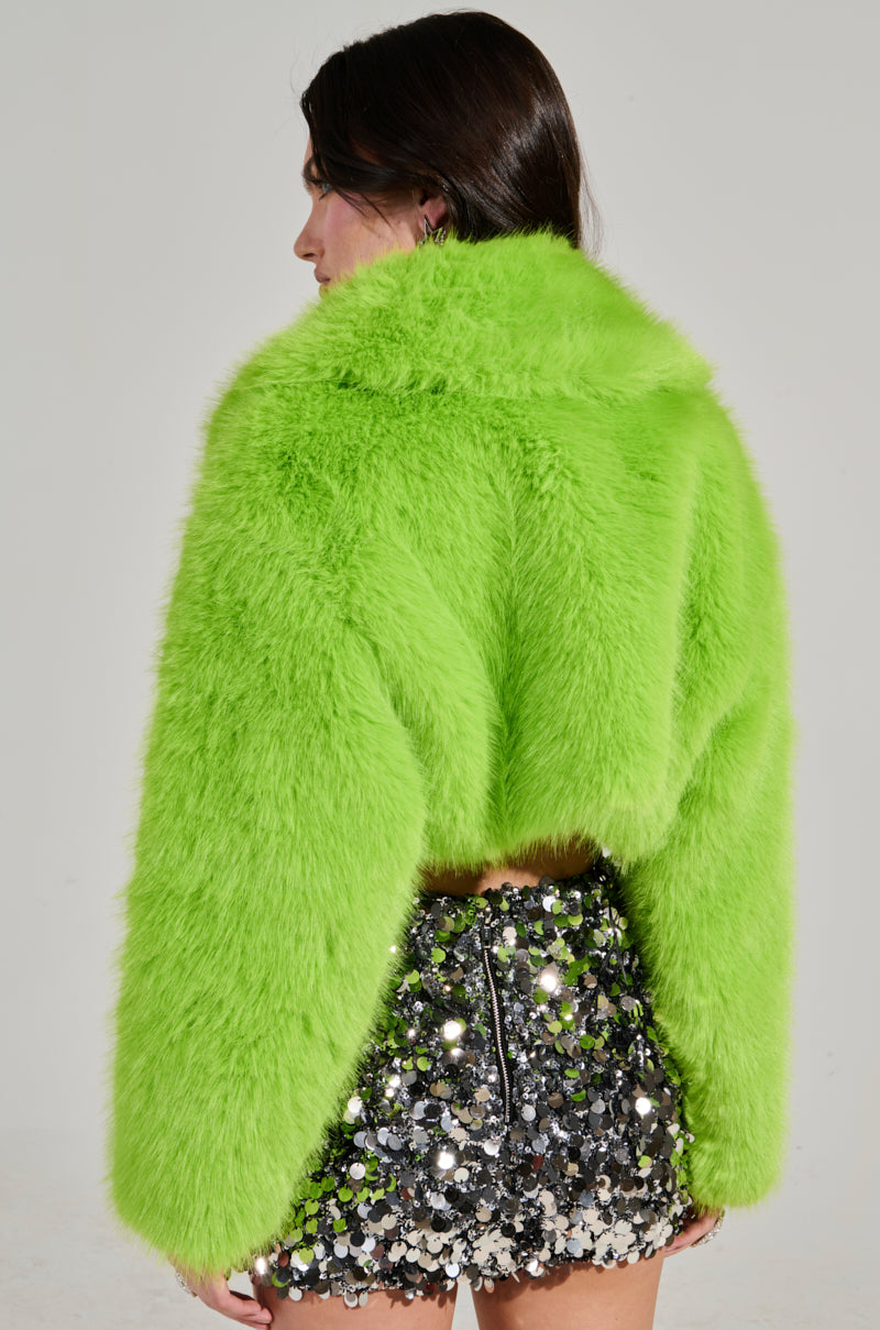 A person with long hair is wearing the vibrant LIGHTS OUT CROPPED FAUX FUR JACKET IN LIME, featuring an oversized collar, paired with a sequined, reflective mini skirt as they stand against a neutral background. The photo captures them from behind.
