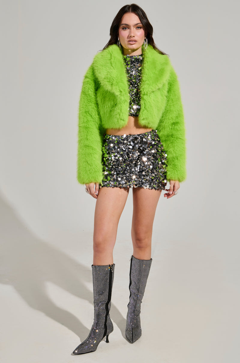 Against a plain backdrop, an individual models the LIGHTS OUT CROPPED FAUX FUR JACKET IN LIME, featuring its distinctive oversized collar. They pair this eye-catching jacket with a sparkly silver top and mini skirt, completing their ensemble with knee-high silver glitter boots.