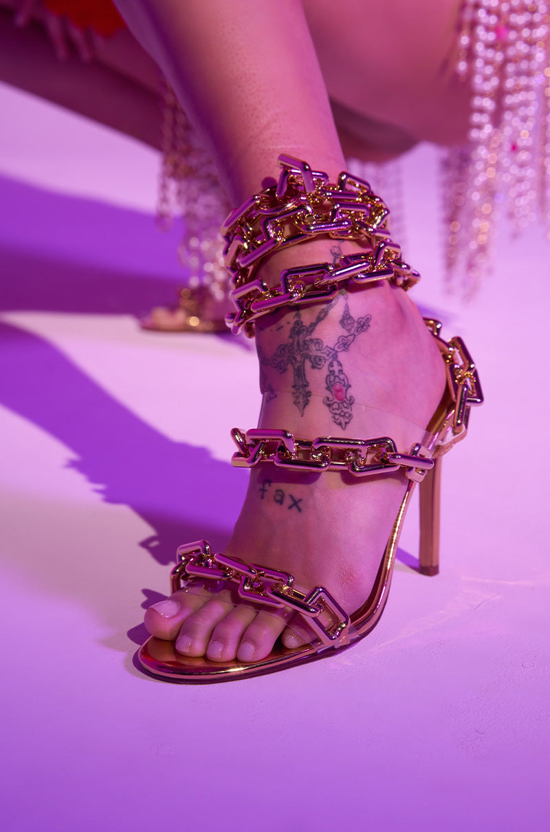 A close-up of a foot wearing the AZALEA WANG YEIRA GOLD SANDAL with chain link details and a stiletto heel. A floral cross tattoo and "fax" are inked on the foot, all set against a purple-toned background.