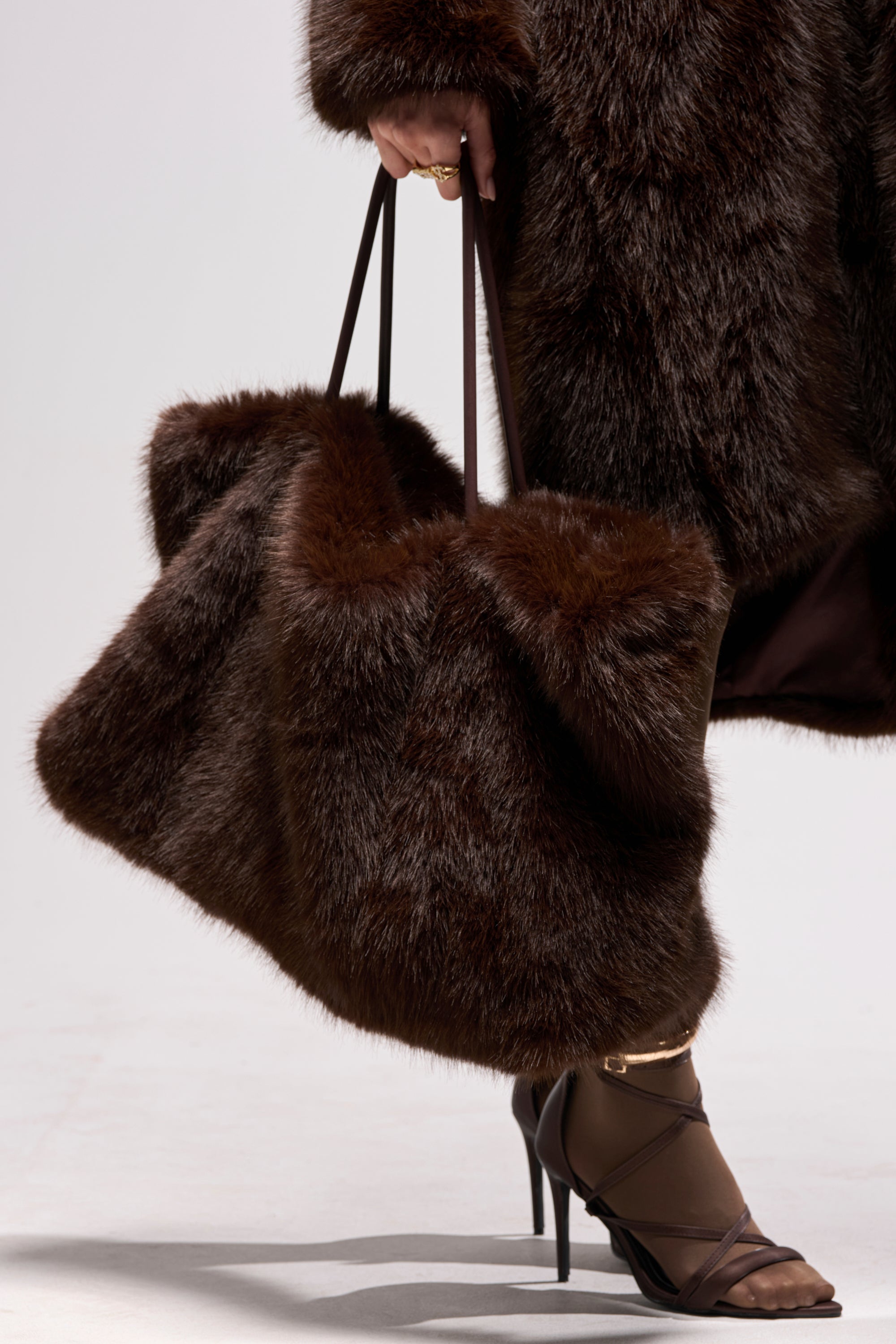 A person in a dark brown fur coat holds the SO RICH FAUX FUR TOTE. They wear brown strappy high-heeled sandals and gold anklets, standing against a plain light background.