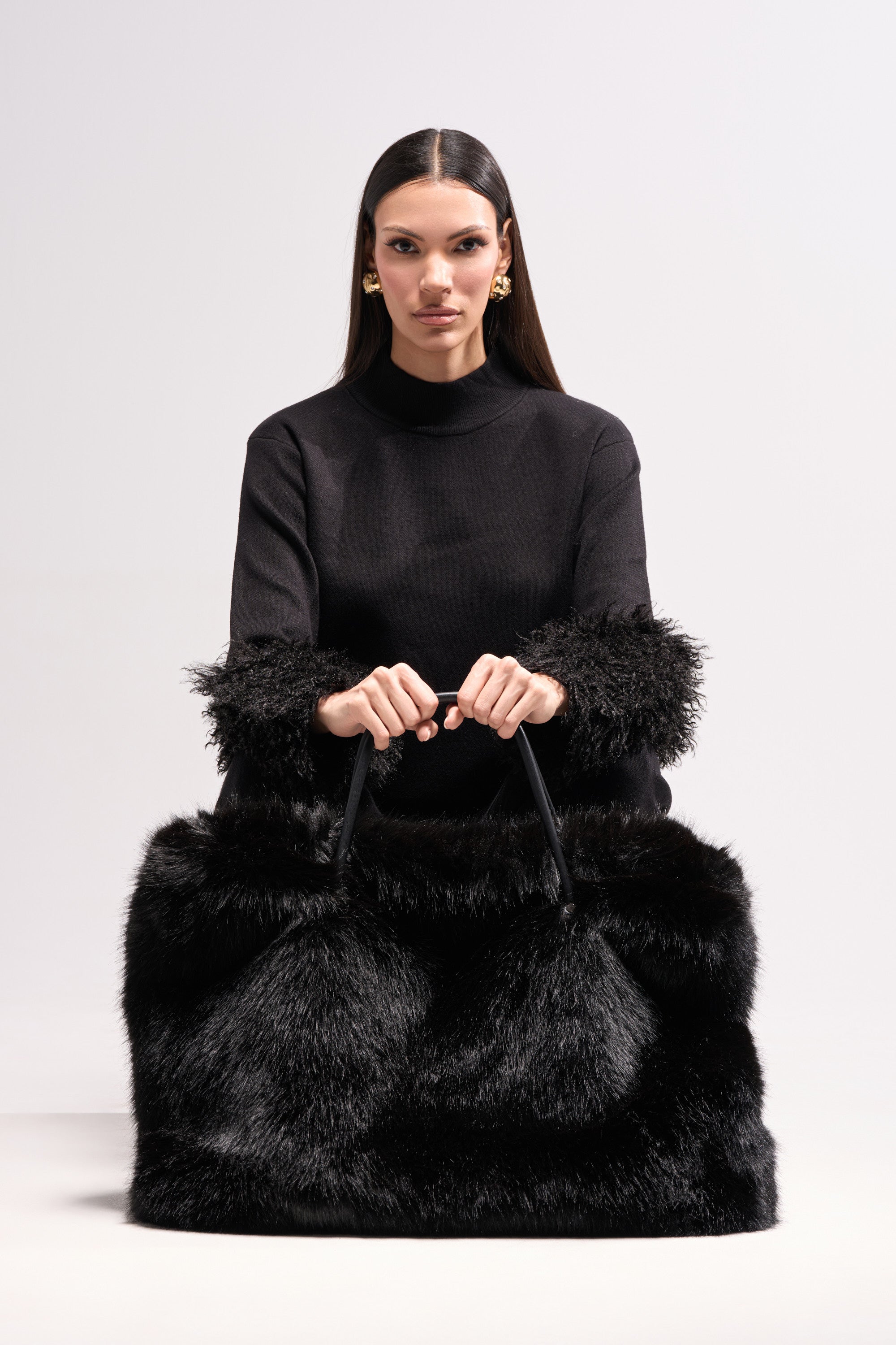 A woman with straight dark hair wears a black top with furry cuffs and holds the YETI LARGE FAUX FUR TOTE IN BLACK, sitting against a plain light background.