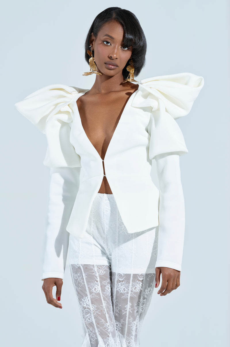 A woman poses in a chic white ensemble featuring the MILA FITTED BLOUSE with a plunging V-neckline and structured puffed sleeves. She completes her look with lace pants, gold earrings, and a sleek hairstyle against a plain background.