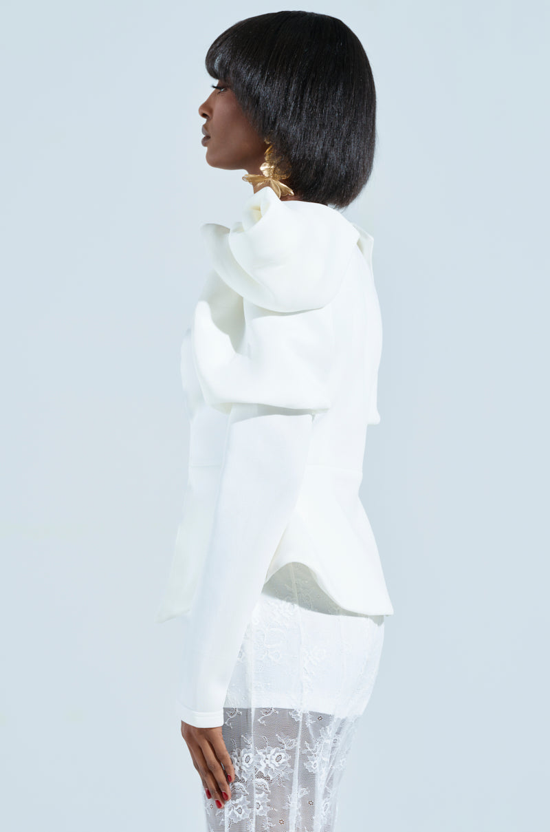 Against a plain backdrop, a person with short hair showcases a stylish white ensemble featuring the MILA FITTED BLOUSE with large shoulder details and a lace skirt. The structured puffed detailing of the outfit adds an elegant touch to their profile.